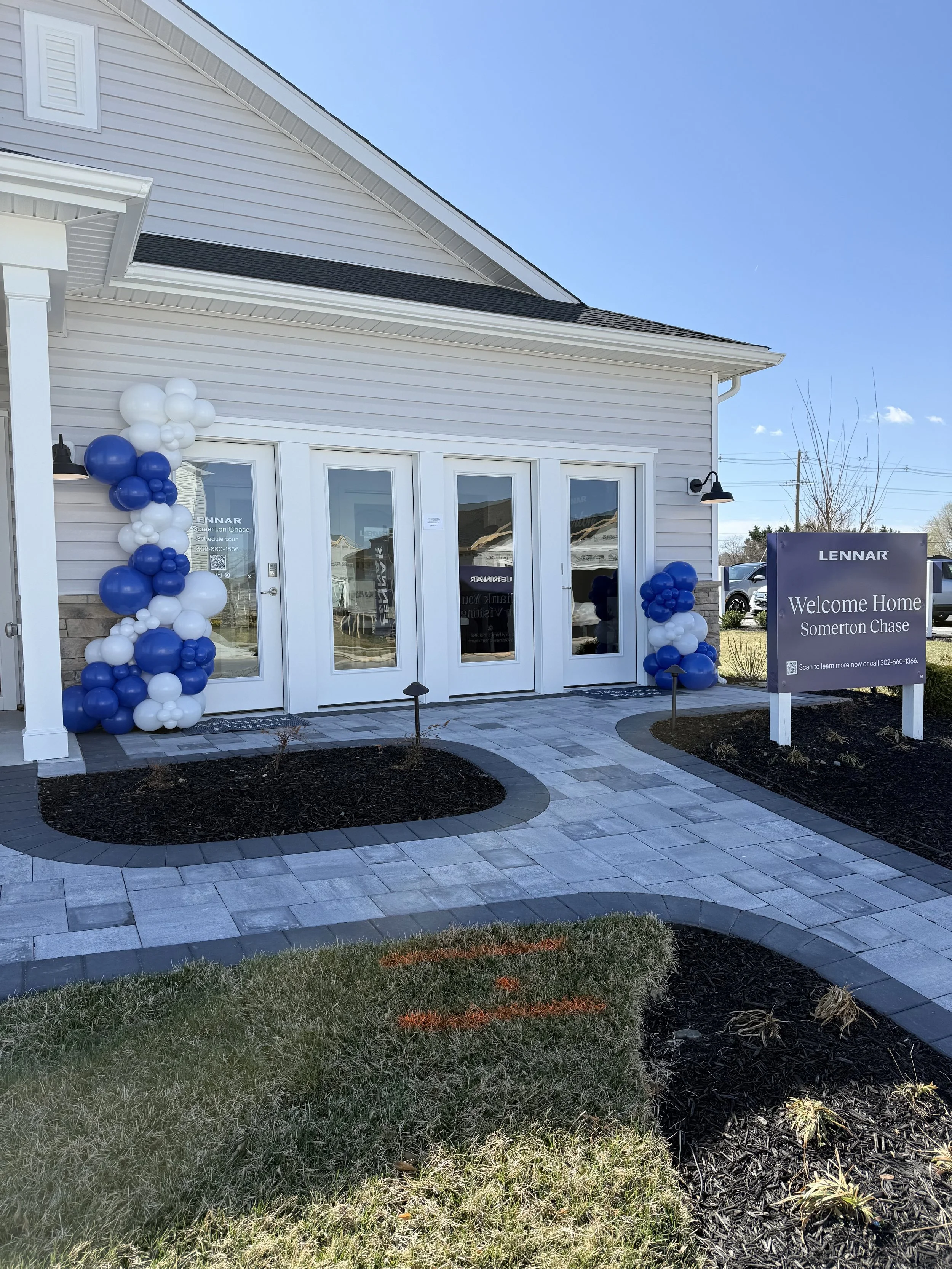 The front of a new house with a porch decorated with blue and white balloons and a sign that reads 'Welcome Home Somerton Chase.' The house has white siding, glass double doors, and outdoor lighting, with a paved walkway and a small landscaped area with grass and young plants.