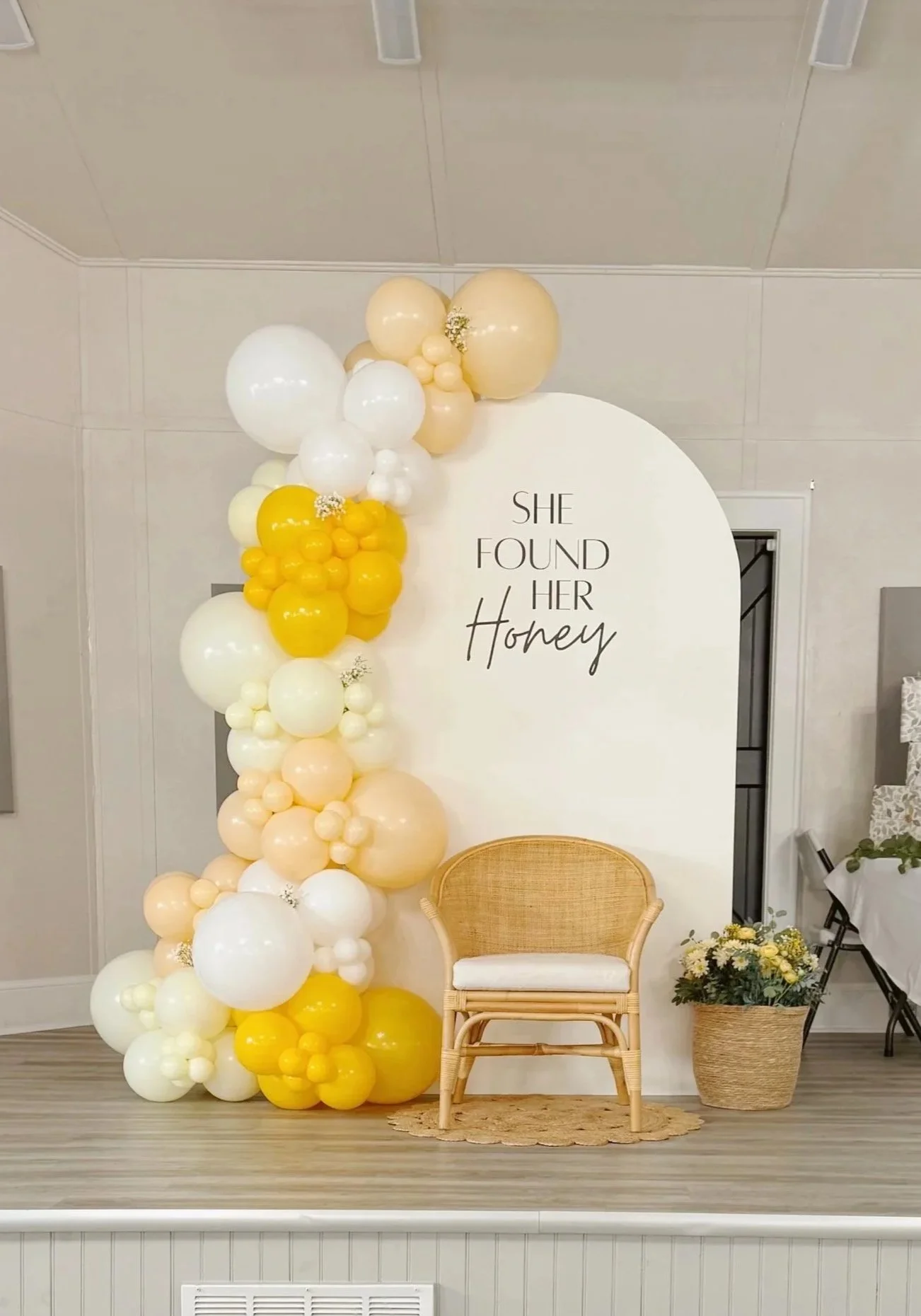 Decorative backdrop with a balloon arrangement in shades of white, yellow, and cream. The backdrop has a sign reading "She Found Her Honey." There is a wicker chair with a white cushion and a basket of flowers next to it on a small woven rug.