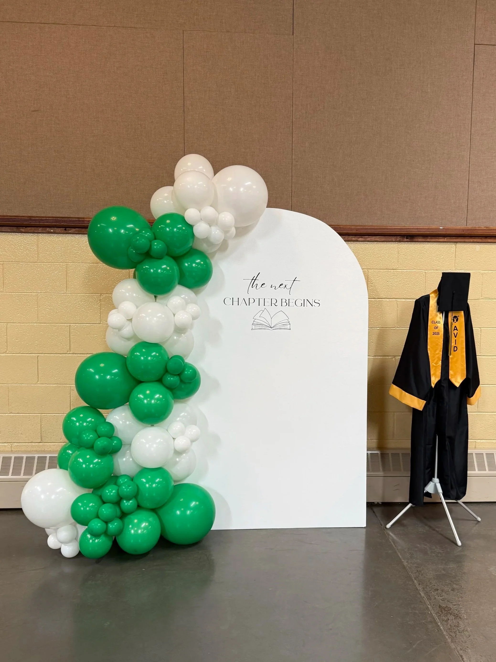 Decorative display with green and white balloons arranged vertically next to a white sign that reads "the next chapter begins" with an open book illustration, and a black graduation gown with a yellow stole hanging on a stand to the right.