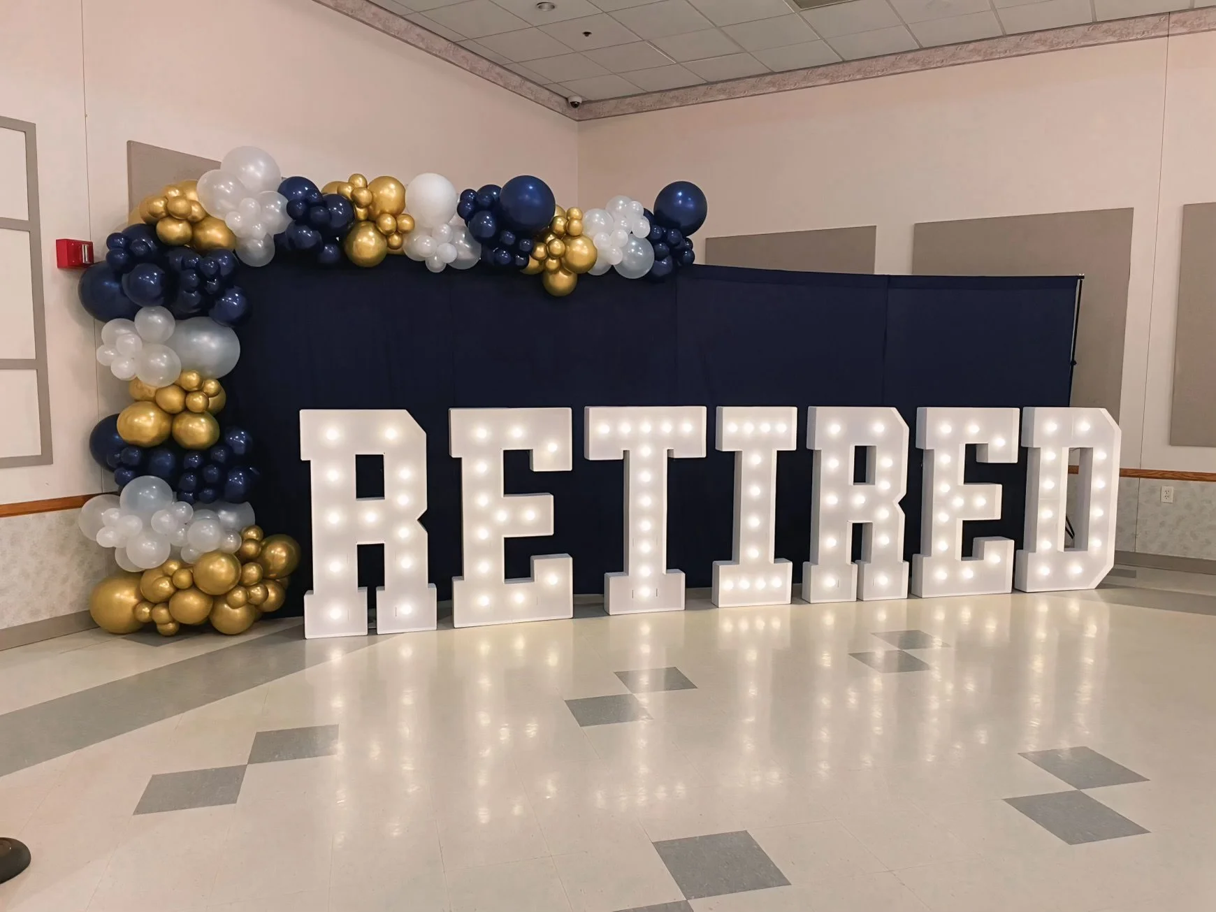 Decorative display with large illuminated letters spelling 'RETRIEVER' and a balloon arrangement in navy blue, white, and gold above and to the side of the letters, set against a dark fabric backdrop in an indoor event space.