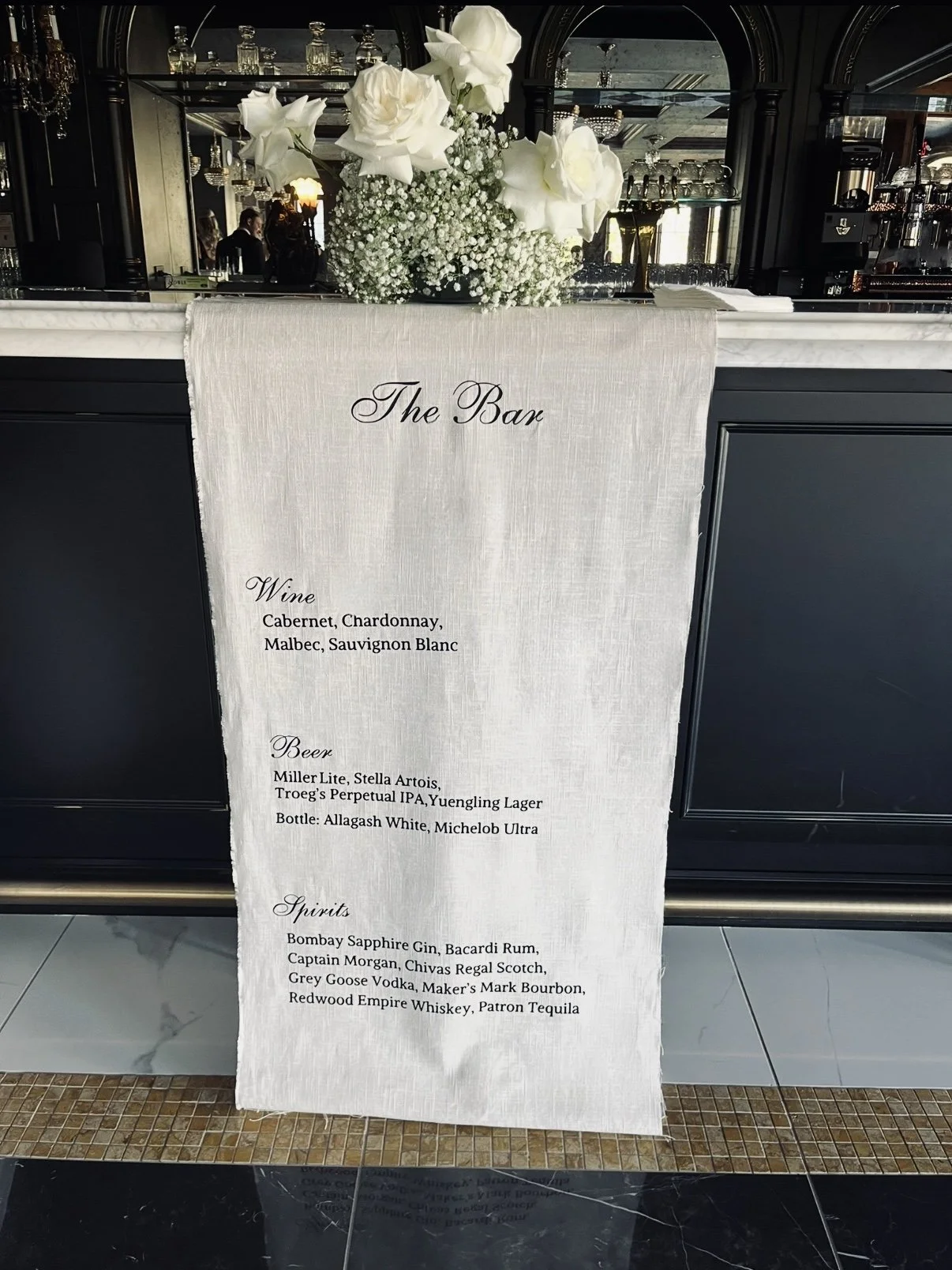 A bar counter with a white cloth sign listing wine, beer, and spirits options, topped with a bouquet of white roses and baby's breath flowers.