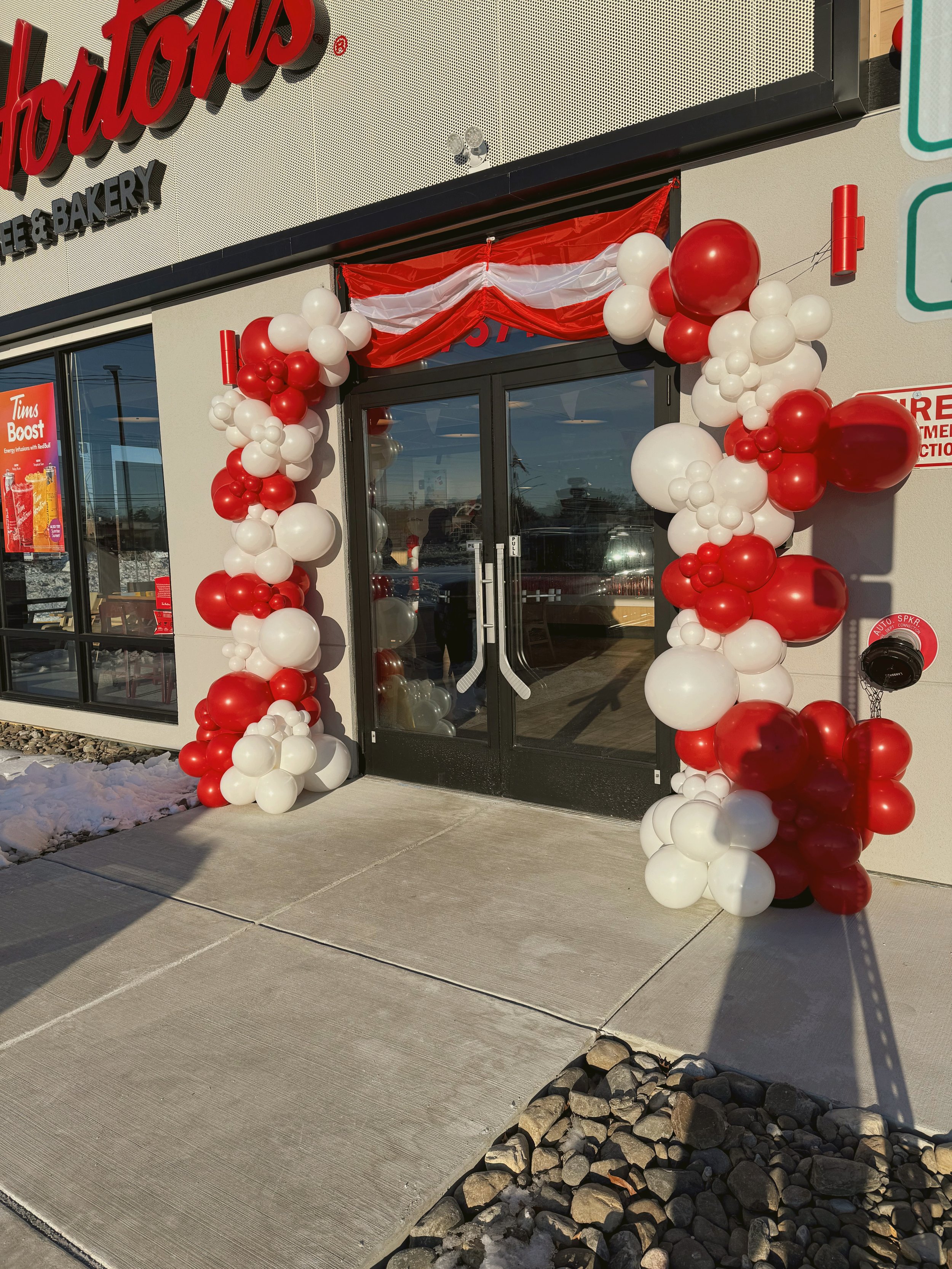Storefront entrance decorated with red and white balloons and red and white fabric drapes, with glass doors and a sign that reads 'Tim Hortons'.