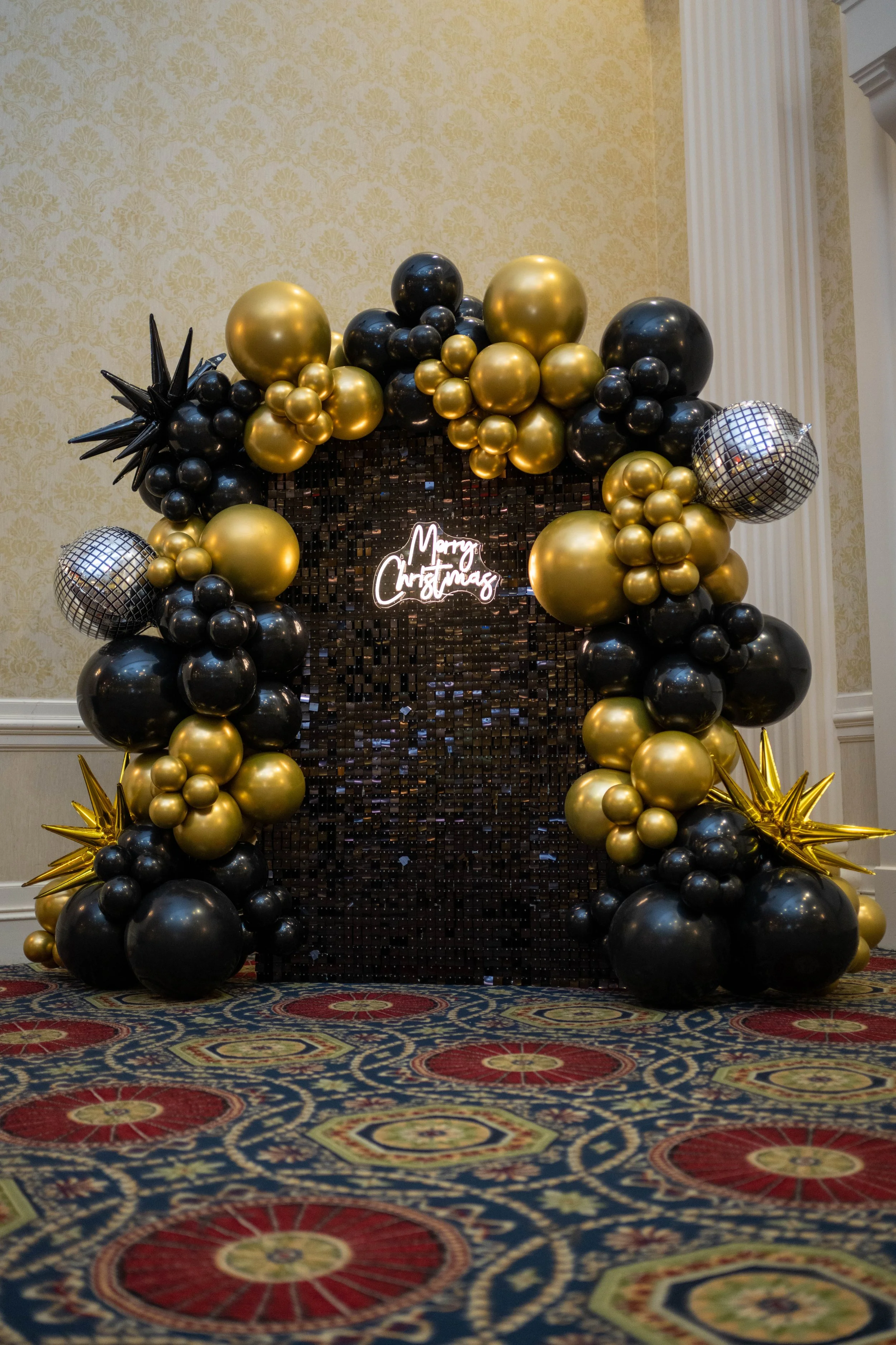 A festive balloon decoration with black, gold, and silver balloons, including star-shaped balloons and disco ball balloons, with a neon sign that says 'Merry Christmas' hanging in the center.