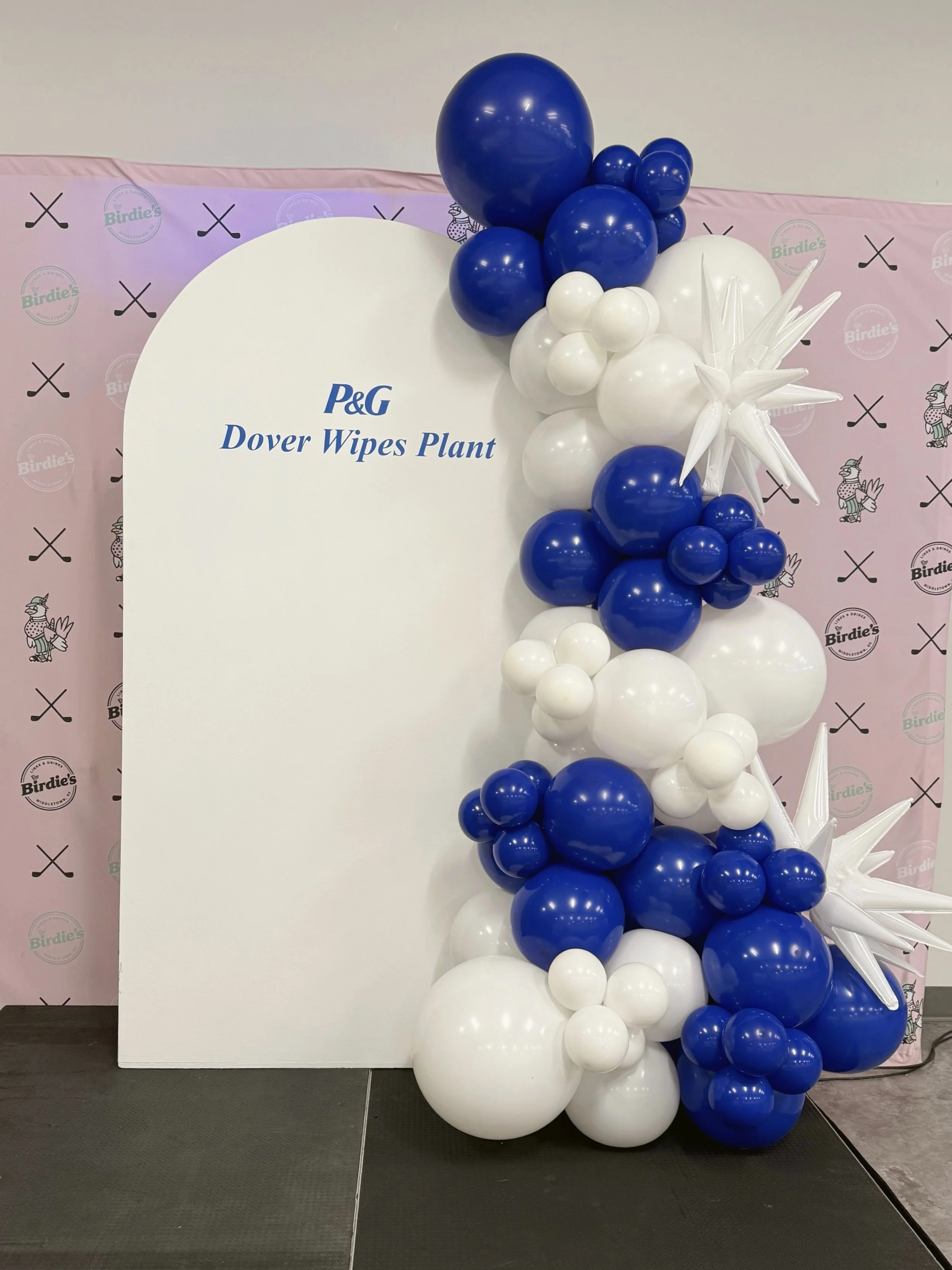 Balloon display with blue and white balloons arranged vertically next to a white board that has the logo and text for P&G Dover Wipes Plant. The background has a pink backdrop with logos and images of cartoon characters.
