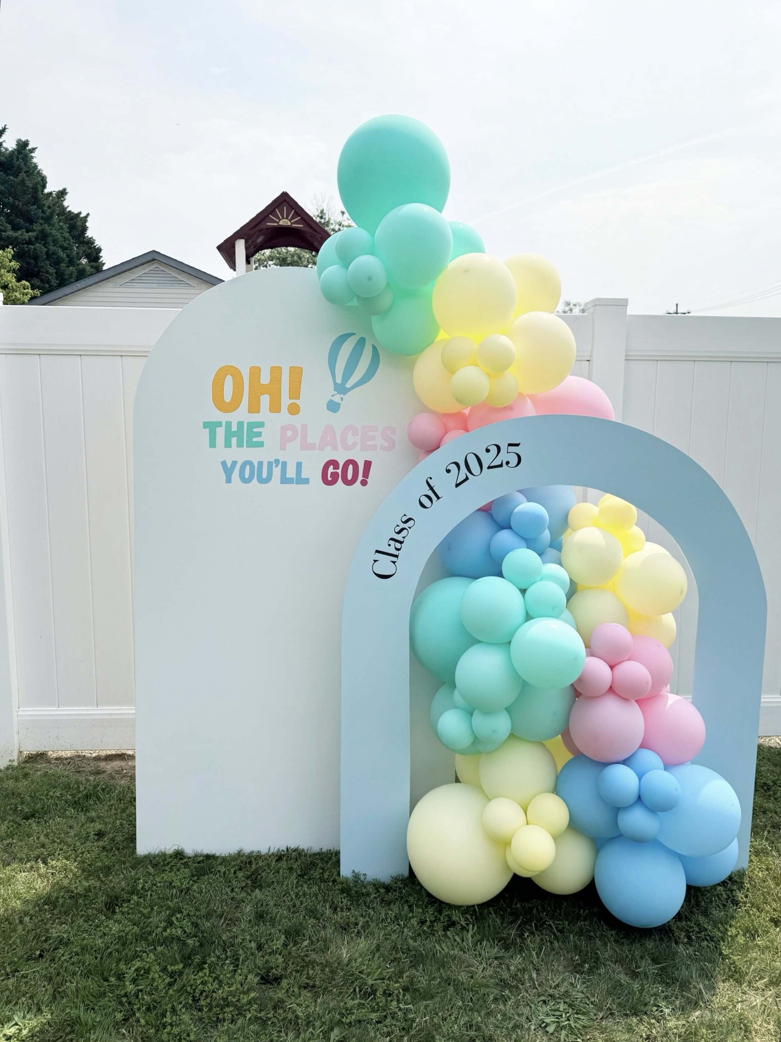Decorative setup with pastel-colored balloons arranged along a white arch and a banner that reads 'OH! THE PLACES YOU'LL GO!' in colorful letters, indicating a graduation celebration.