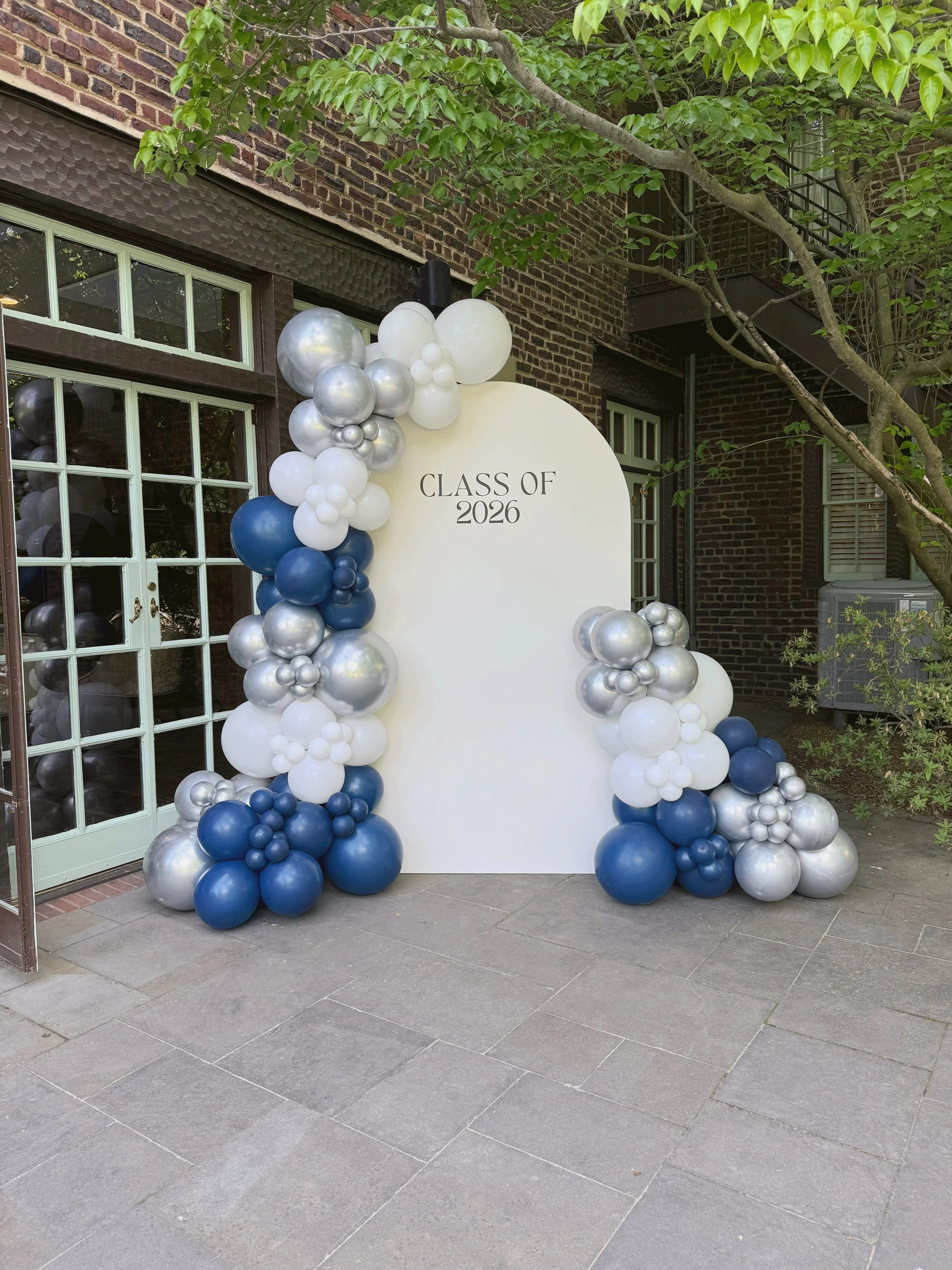 A graduation celebration decoration with a white backdrop and balloons in silver, white, and navy blue. The backdrop has the text "Class of 2026" written on it. The scene is set outside near a brick building with a window and surrounding greenery.