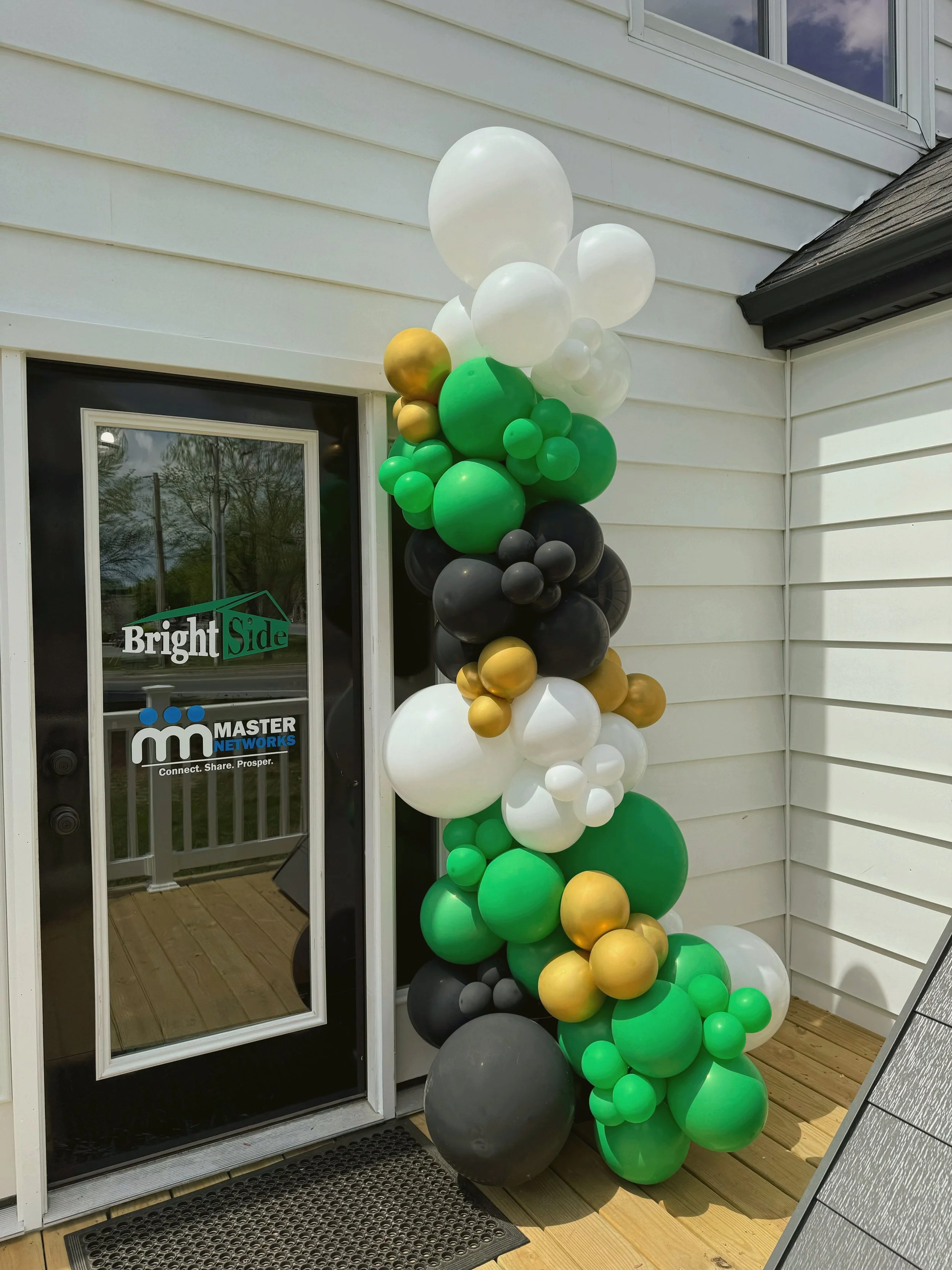 A balloon arrangement with green, white, black, gold, and gray balloons decorate the outside of a house by the door.