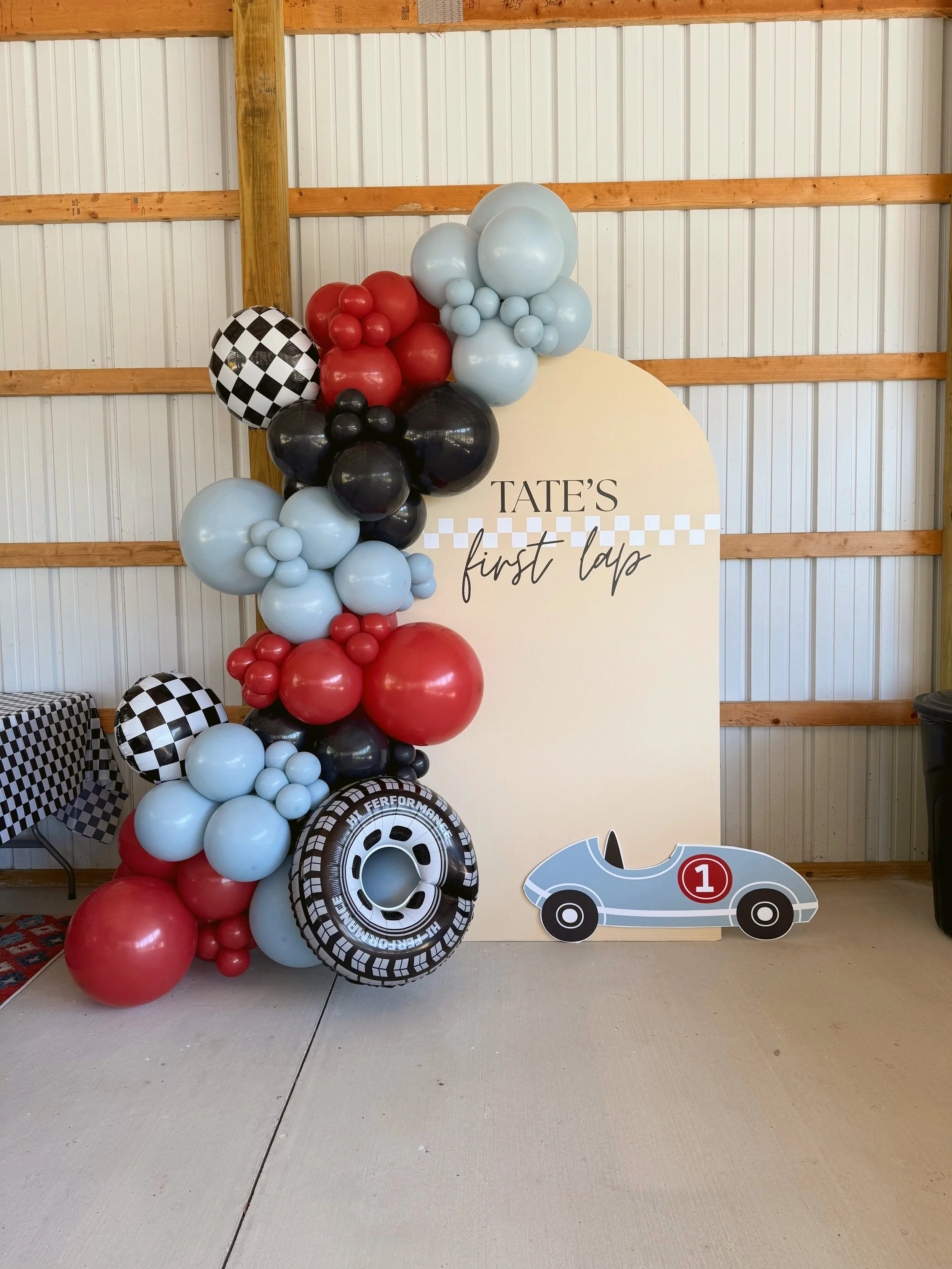 Decorative display with black, red, and light blue balloons, a car-themed cutout with the number 1, and a sign that reads "Tate's first lap" against a metal-paneled wall.