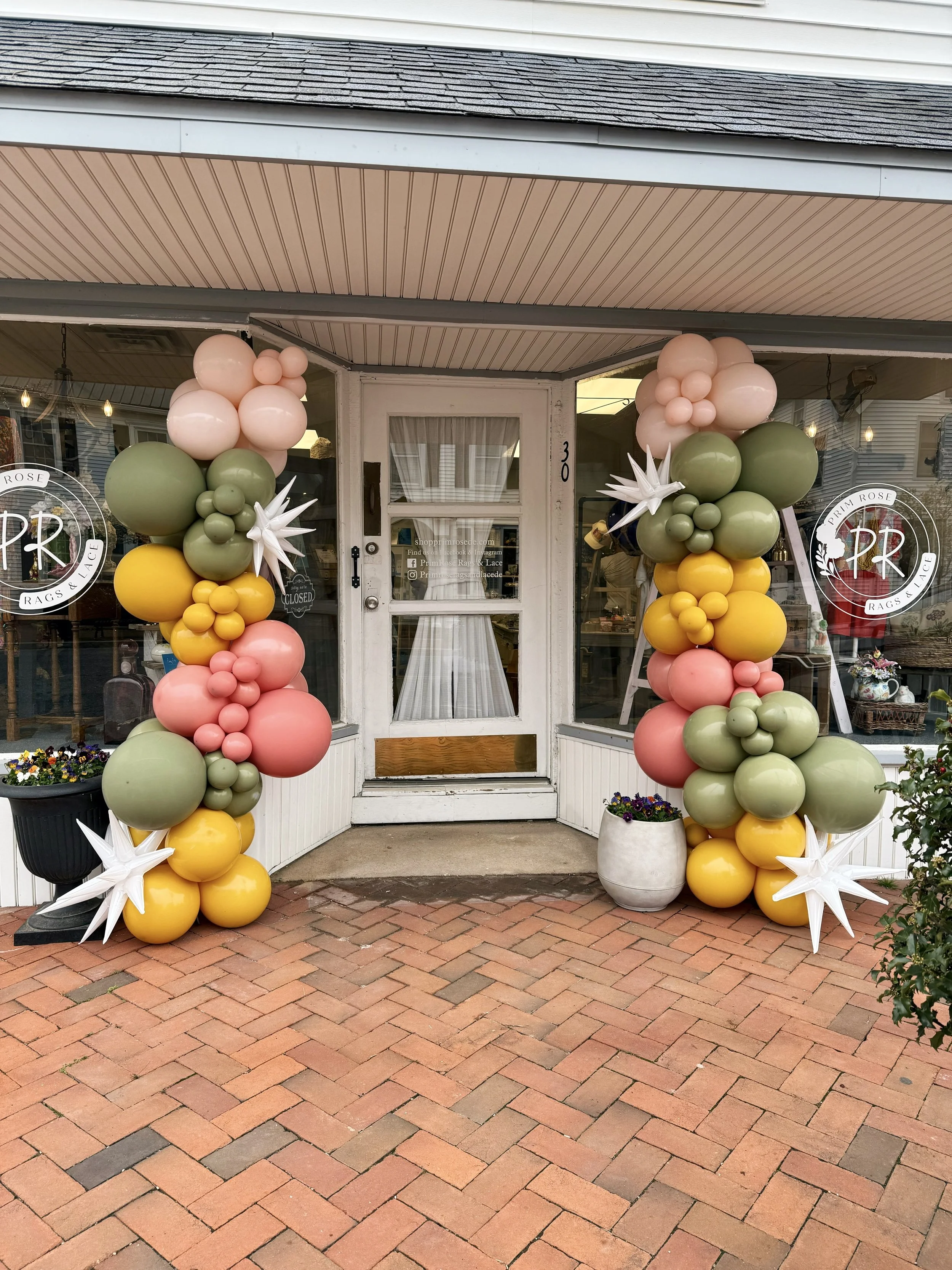 Decorative balloon arch with pastel pink, green, yellow, and white balloons, accented with white star-shaped balloons, placed in front of a storefront with glass doors, flanking the entrance on both sides.