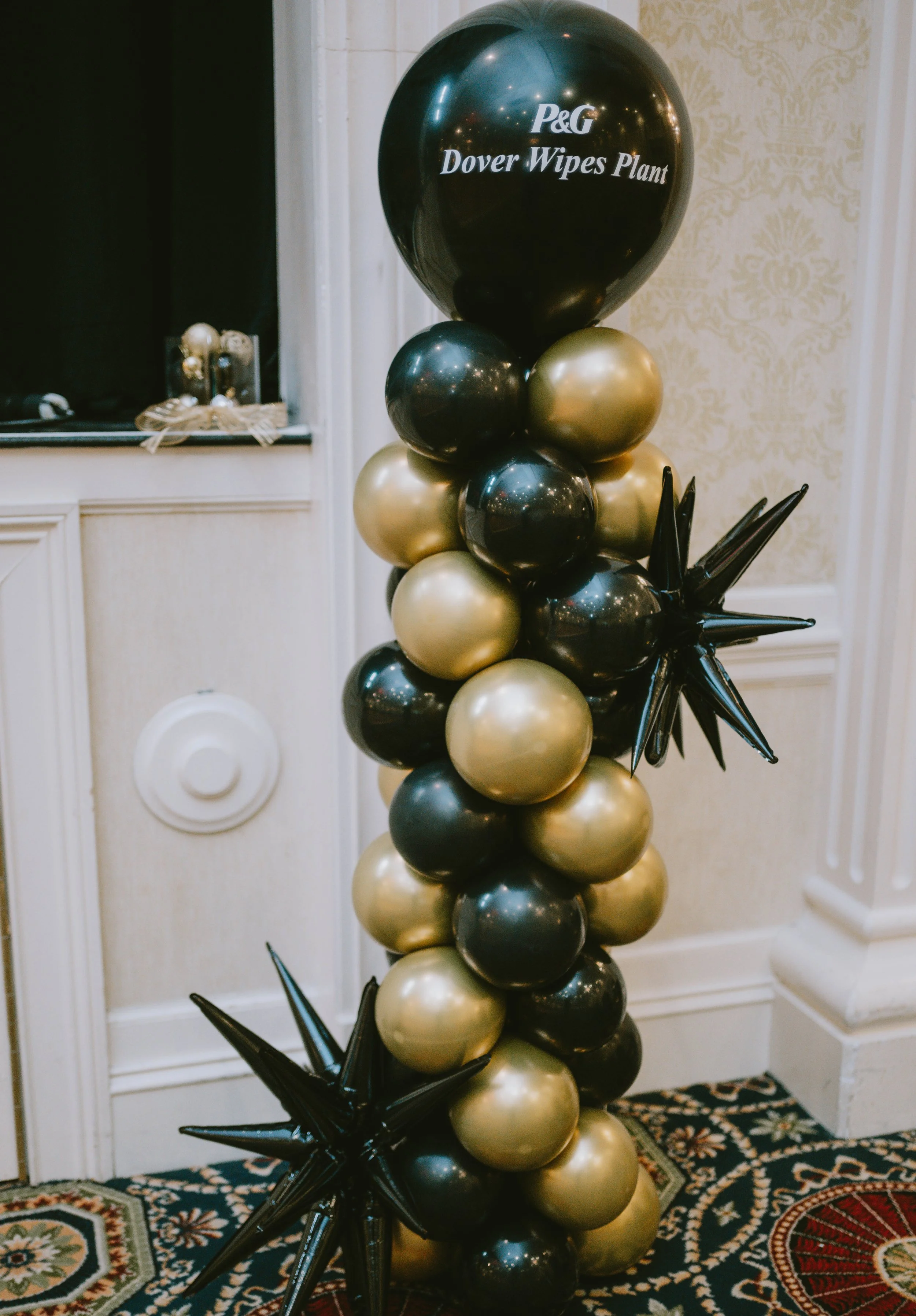 Black and gold balloons arranged in a column with black star-shaped balloons at the base. The balloon at the top reads 'P&G Dover Wipes Plant'.