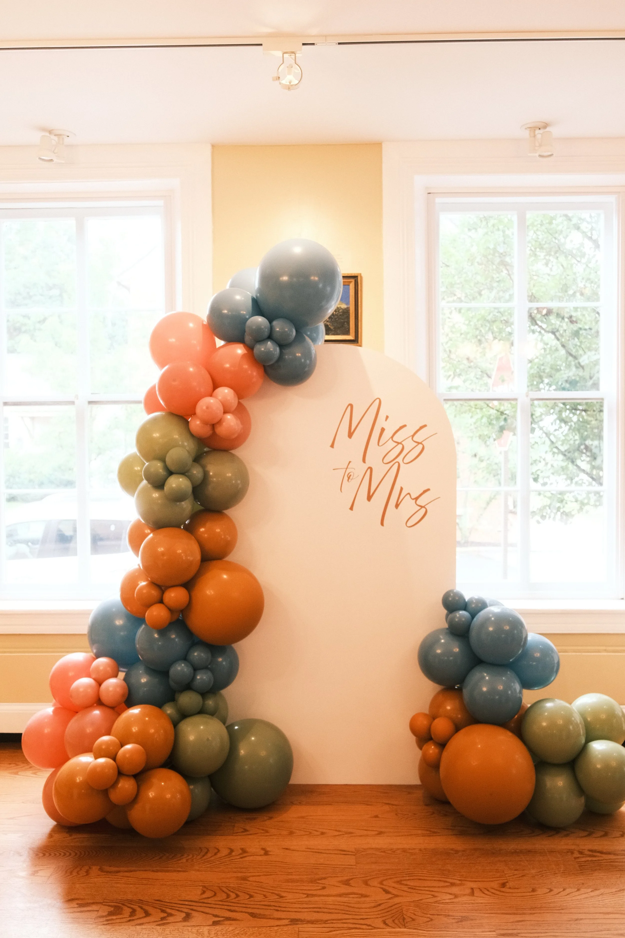 Decorative balloon arrangement with orange, sage green, and light blue balloons around a white backdrop that has 'Miss to Mrs' written on it, set inside a bright room with large windows.