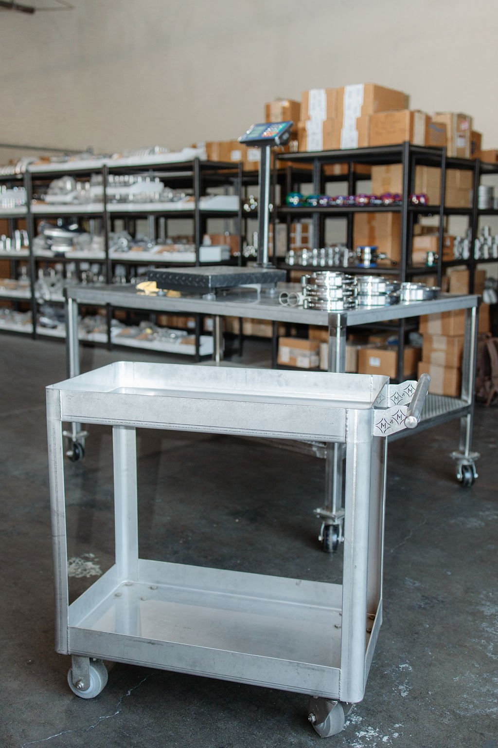 Metal rolling cart in front of shelves filled with hardware parts and boxes in a warehouse or workshop.