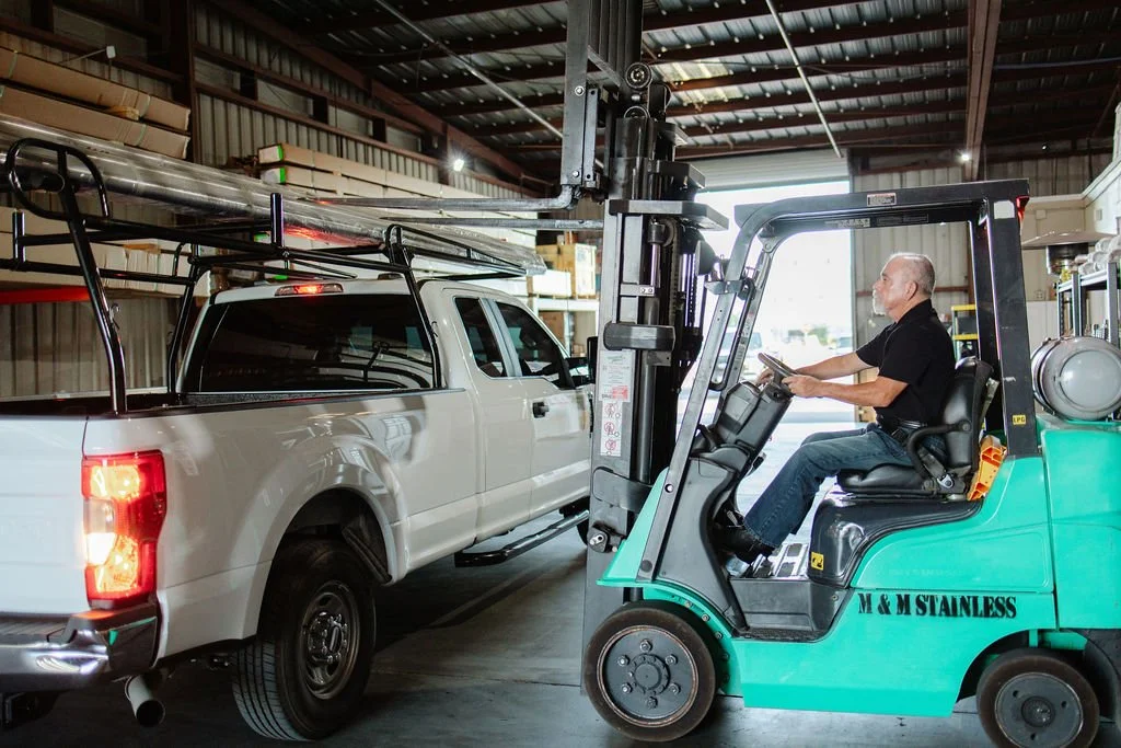 A man operating a green forklift inside a warehouse, moving a white pickup truck with ladders on its roof.