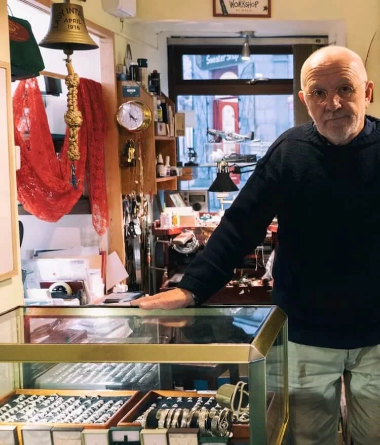 A man wearing a black sweater and tan slacks standing inside a stor with his hand on a glass jewelry case