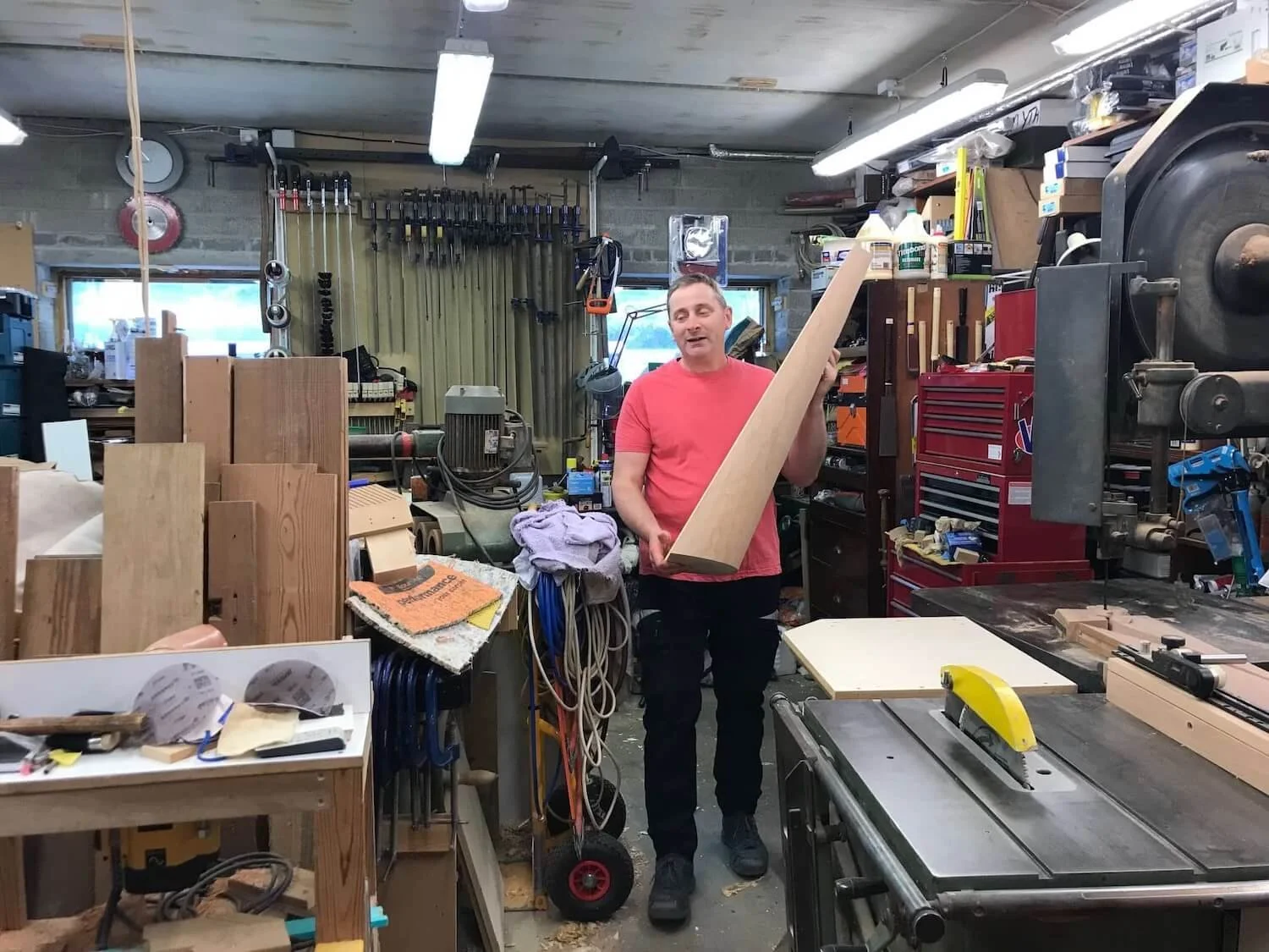 Man holding a piece of wood surrounded by wood and tools in the harp workshop
