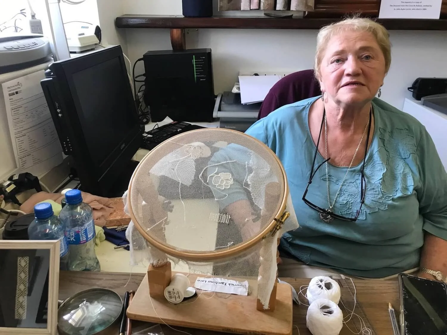a woman lacemaker wearing a blue top and sitting next to her lace project in progress