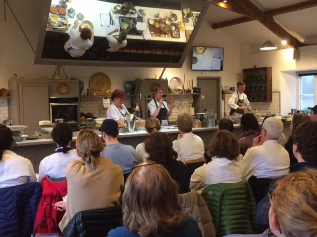back of a group of people watching three chefs in a kitchen perform a cooking demonstration