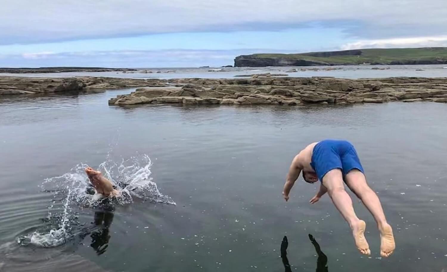 Two people diving into calm, rocky coastal water; one mid-air, the other splashing into the surface.