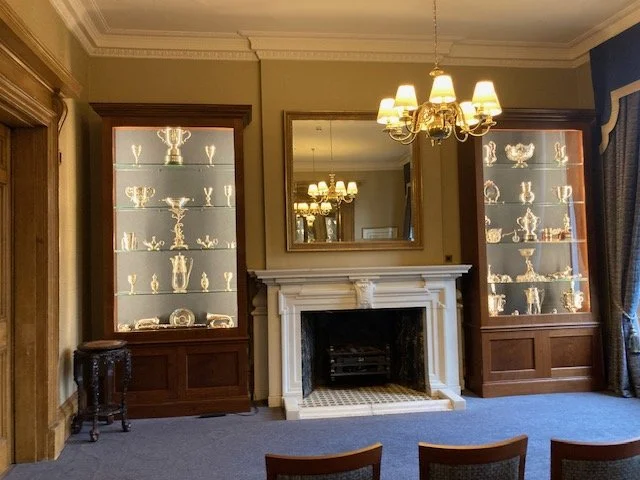 a room with chairs, a fireplace, blue carpet and military silver displayed in wood and glass cabinets flanking the fireplace