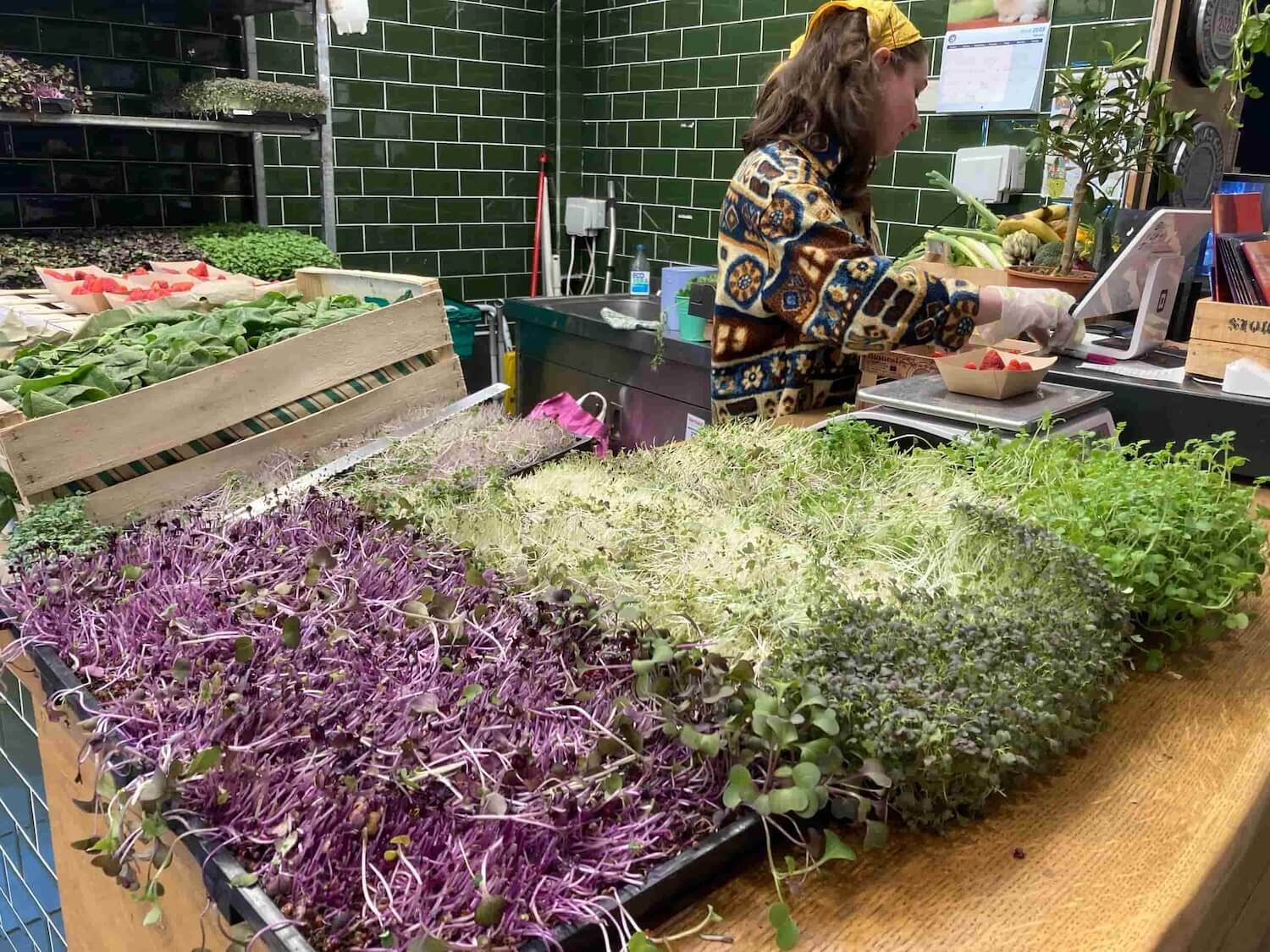 herb vendor weighing strawberries. wooden bins of herbs in the foreground and green subway tile walls in the background