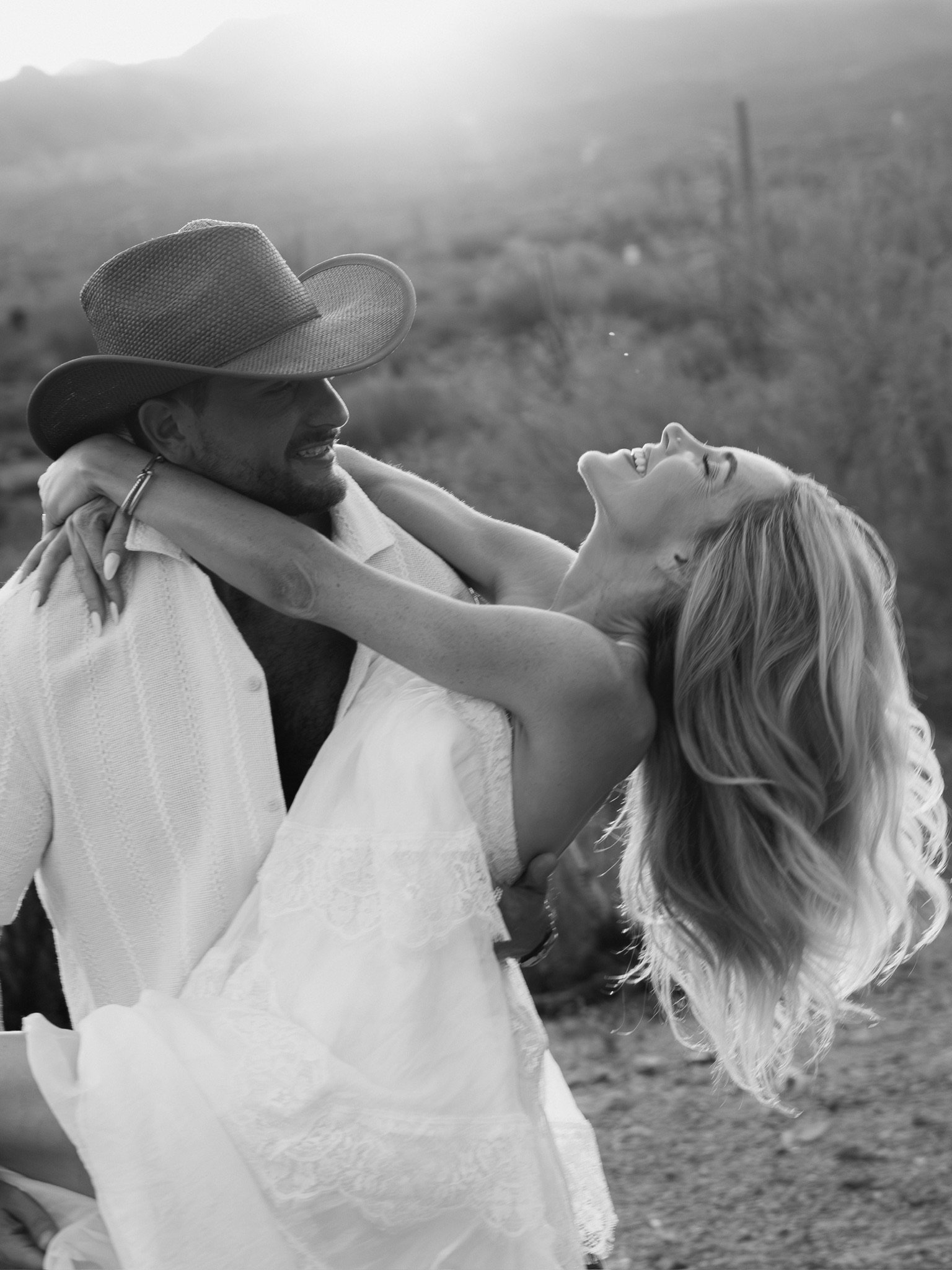A man wearing a cowboy hat and a woman with long hair are smiling and looking at each other, holding each other closely outdoors in Scottsdale Arizona