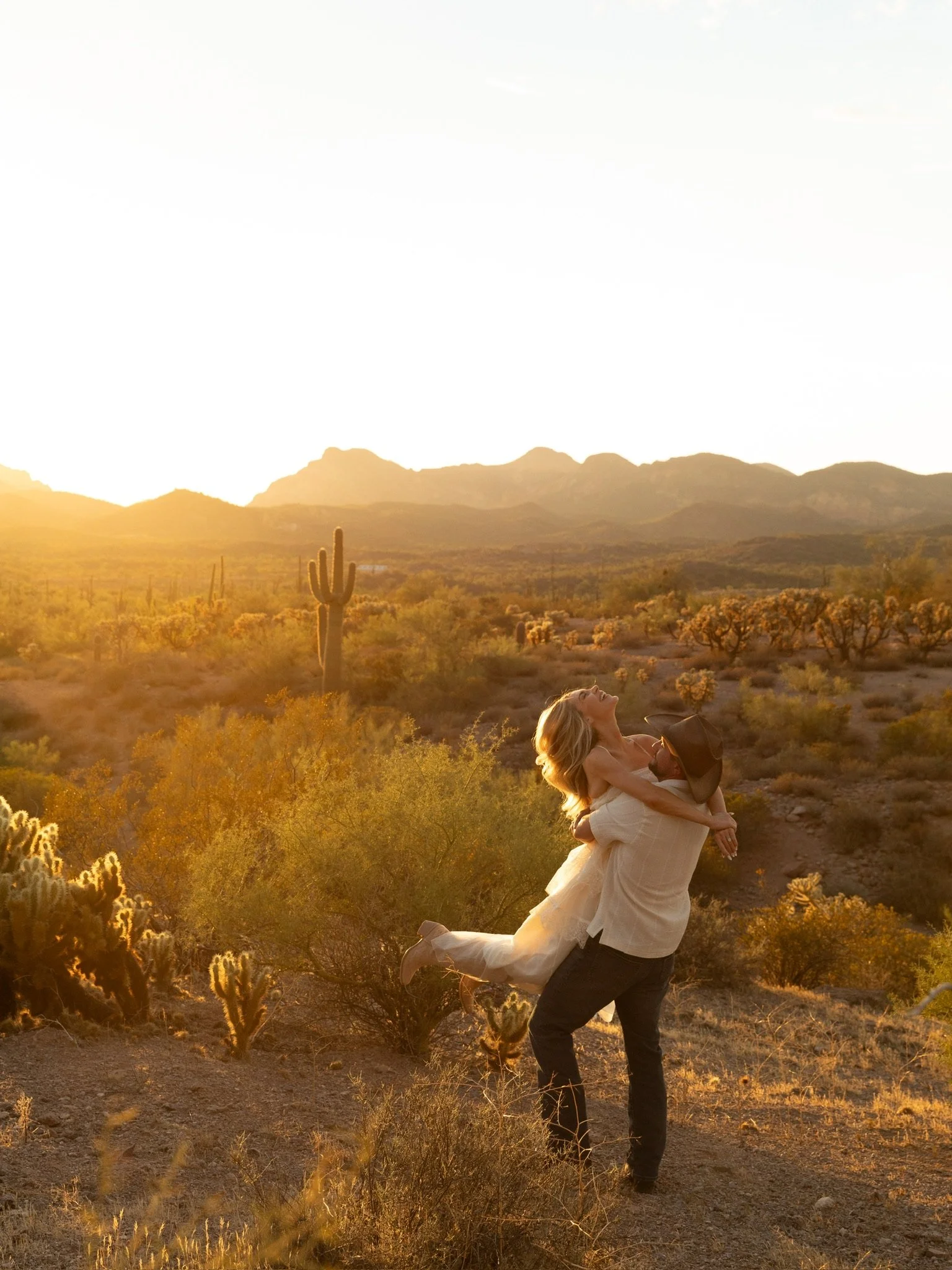 A man in dark pants and a white shirt is lifting a woman in a white dress in the air in a desert landscape with cacti and mountains in the background during sunset.