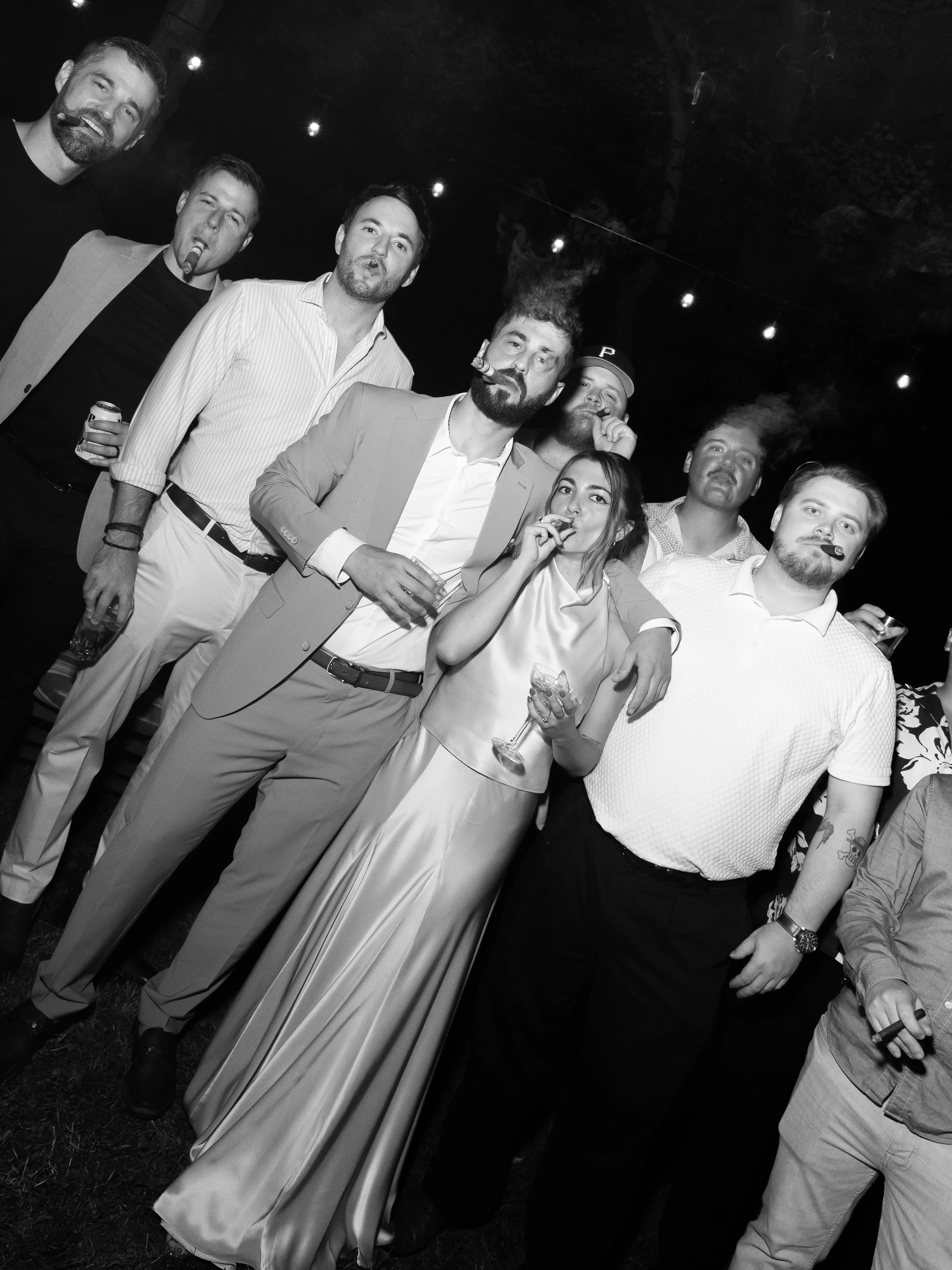 A group of seven people, five men and two women, dressed in formal attire at an outdoor night event. They are smoking cigars and holding drinks, with some making playful facial expressions. String lights are visible overhead.