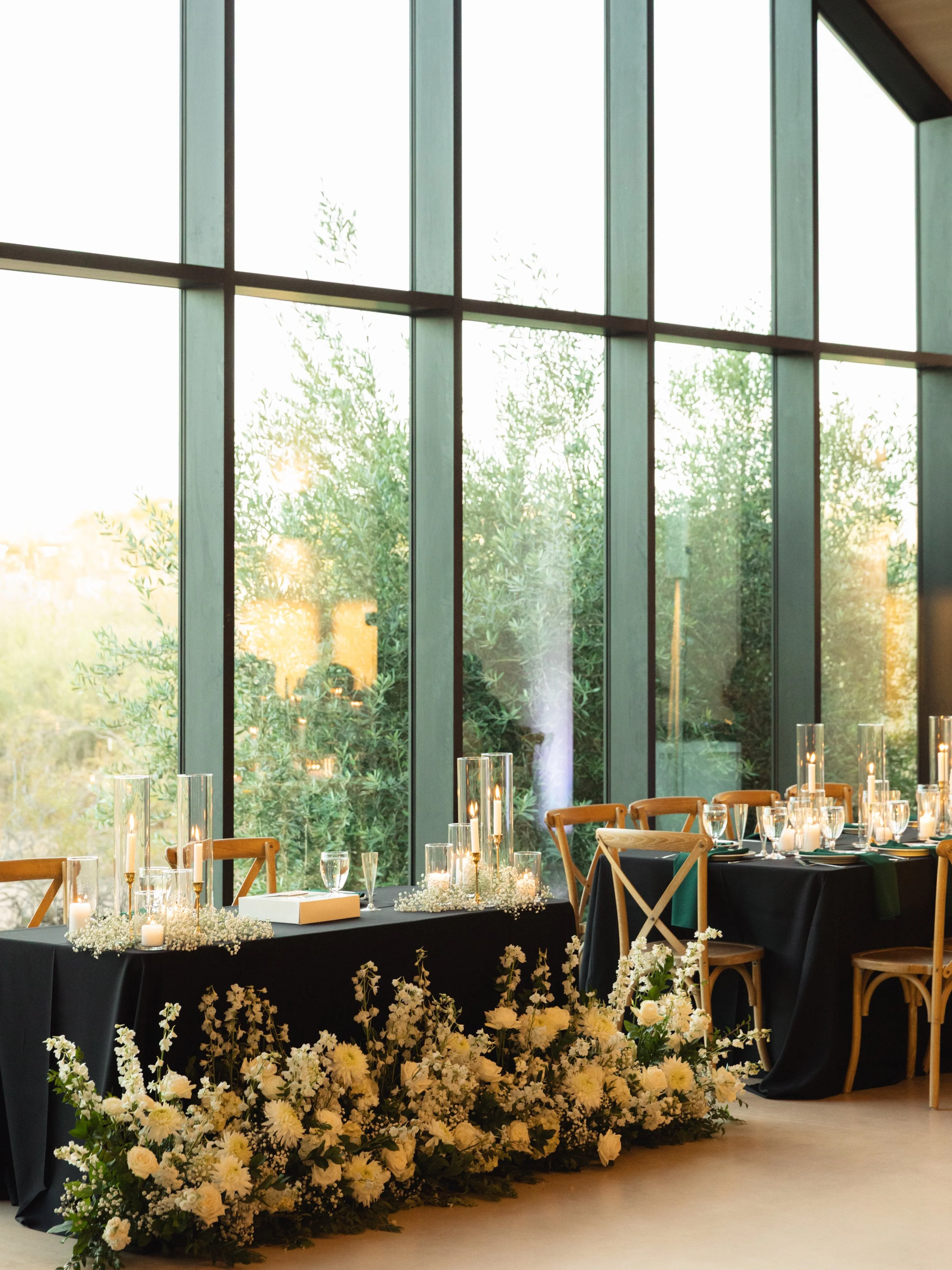 Wedding reception area with a large floral arrangement of white flowers and candles on a black-draped table, set in front of tall floor-to-ceiling windows with trees visible outside.
