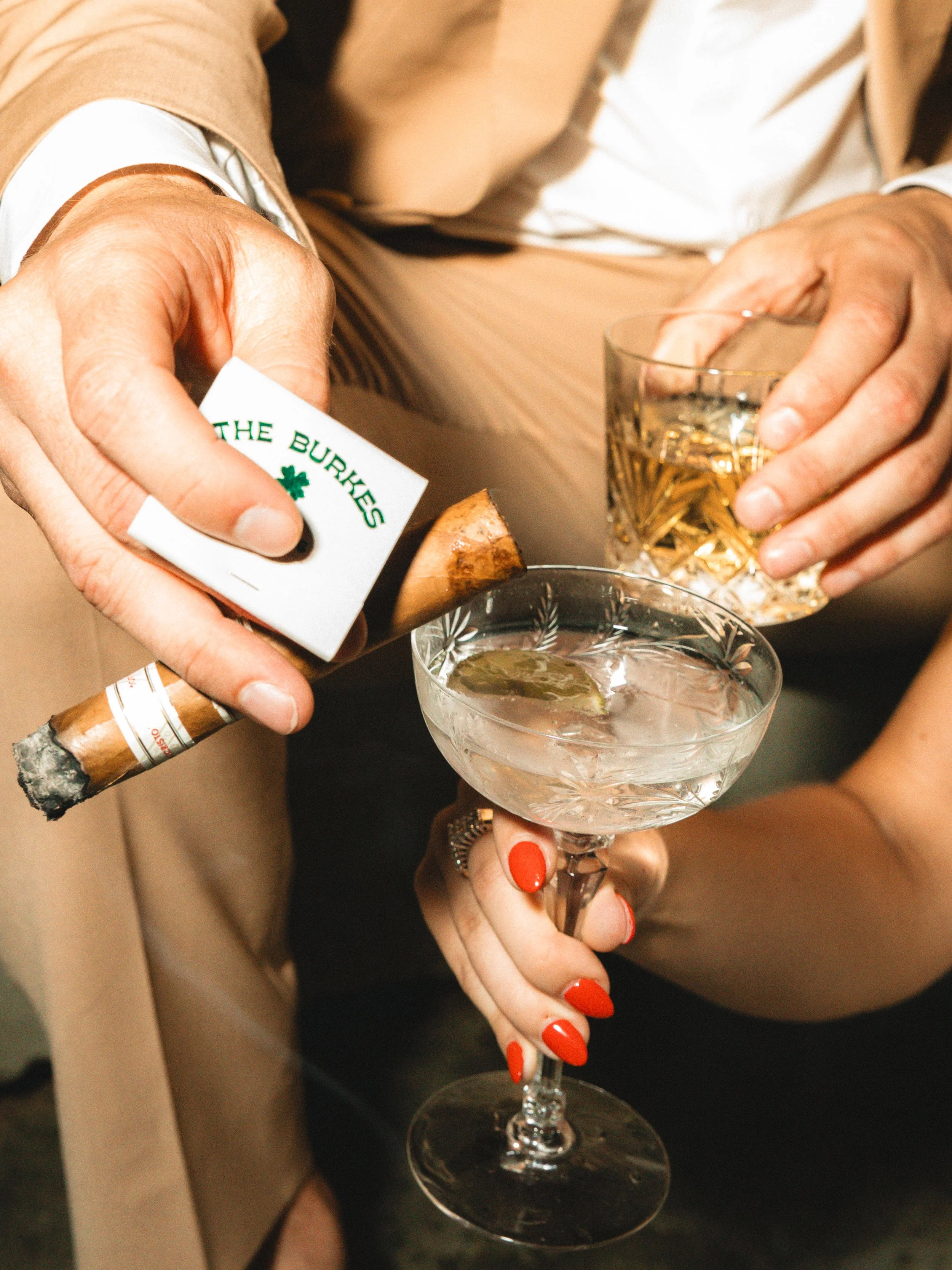 A person with red nails holding a cocktail glass with a slice of lime, into which a hand with a cigar is pouring a drink from a bottle. The person is wearing a black dress and a beige blazer.