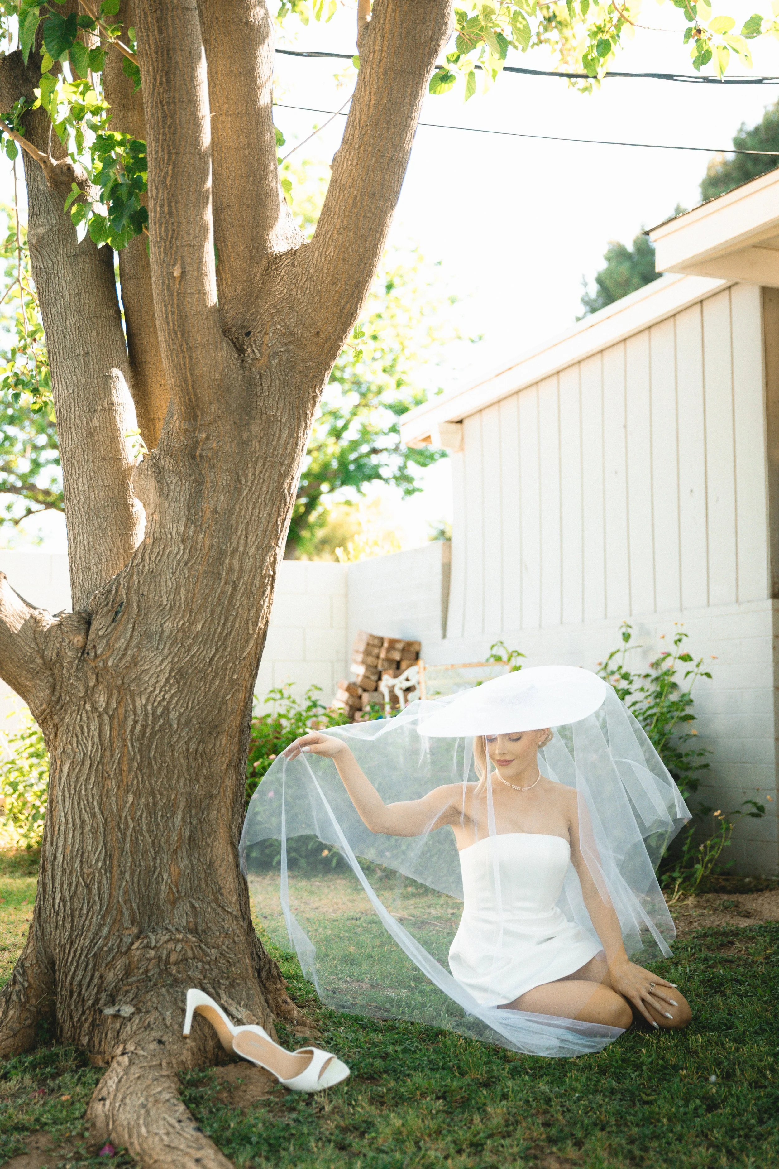 A woman in a wedding dress sits on the grass beneath a tree, holding a sheer veil. A white high-heeled shoe is on a branch near her. She is wearing a pearl necklace, and her hair is styled up. In the background, there is a white building, some stacke