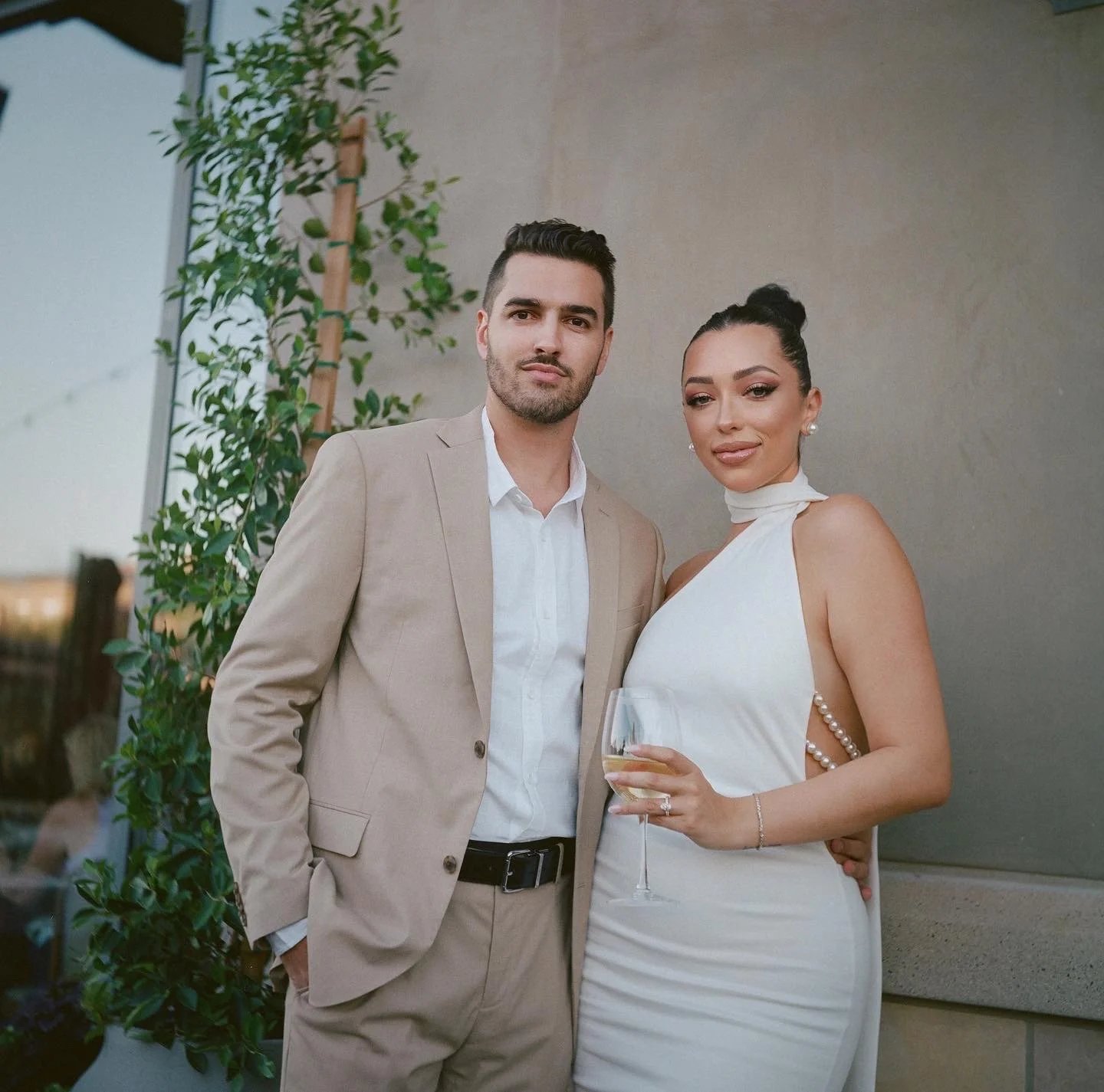 A man in a beige suit and a woman in a white dress holding a glass of white wine at a social event.