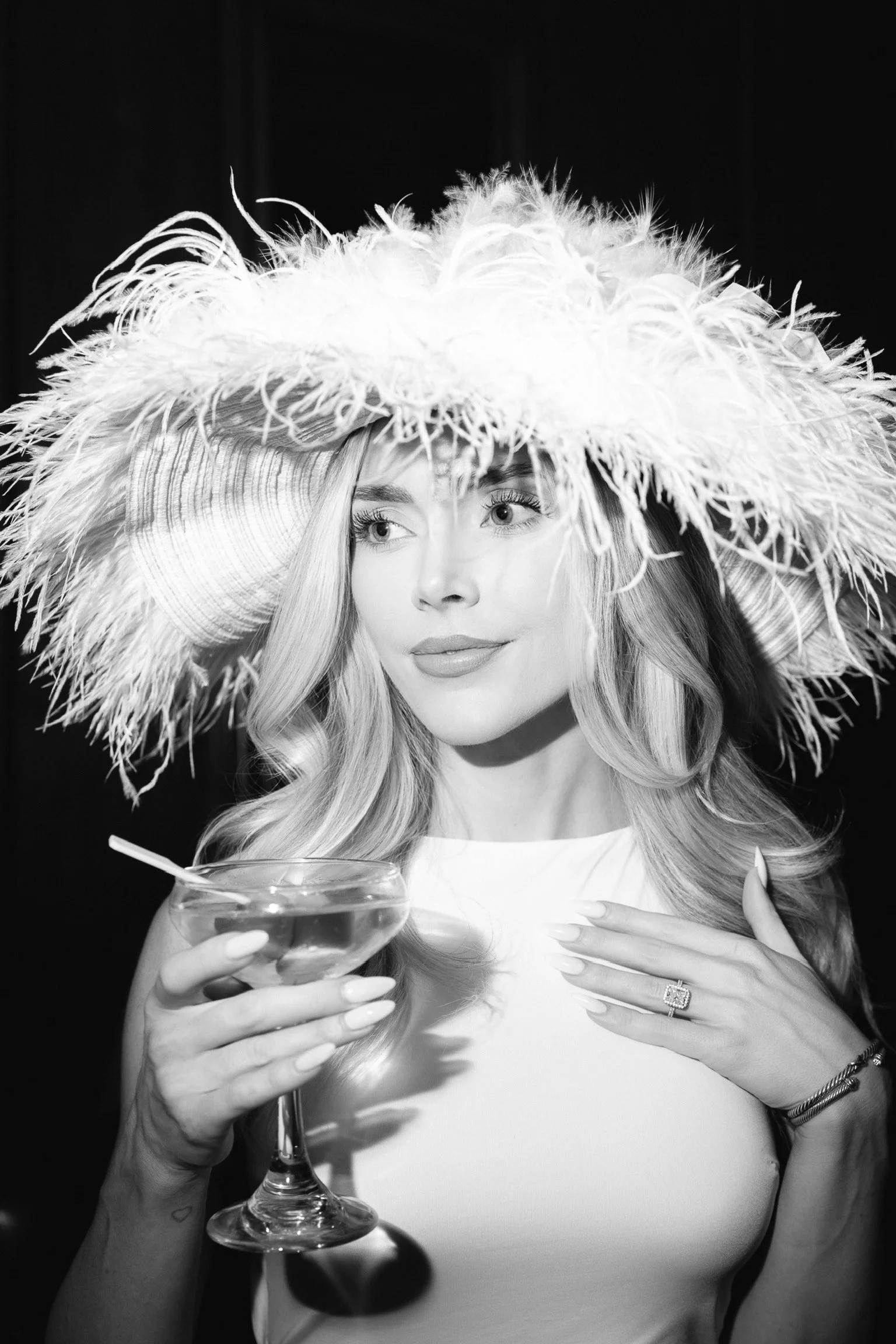 Black and white photo of a woman with long, wavy hair, wearing a feathered hat, holding a cocktail glass, and smiling softly.
