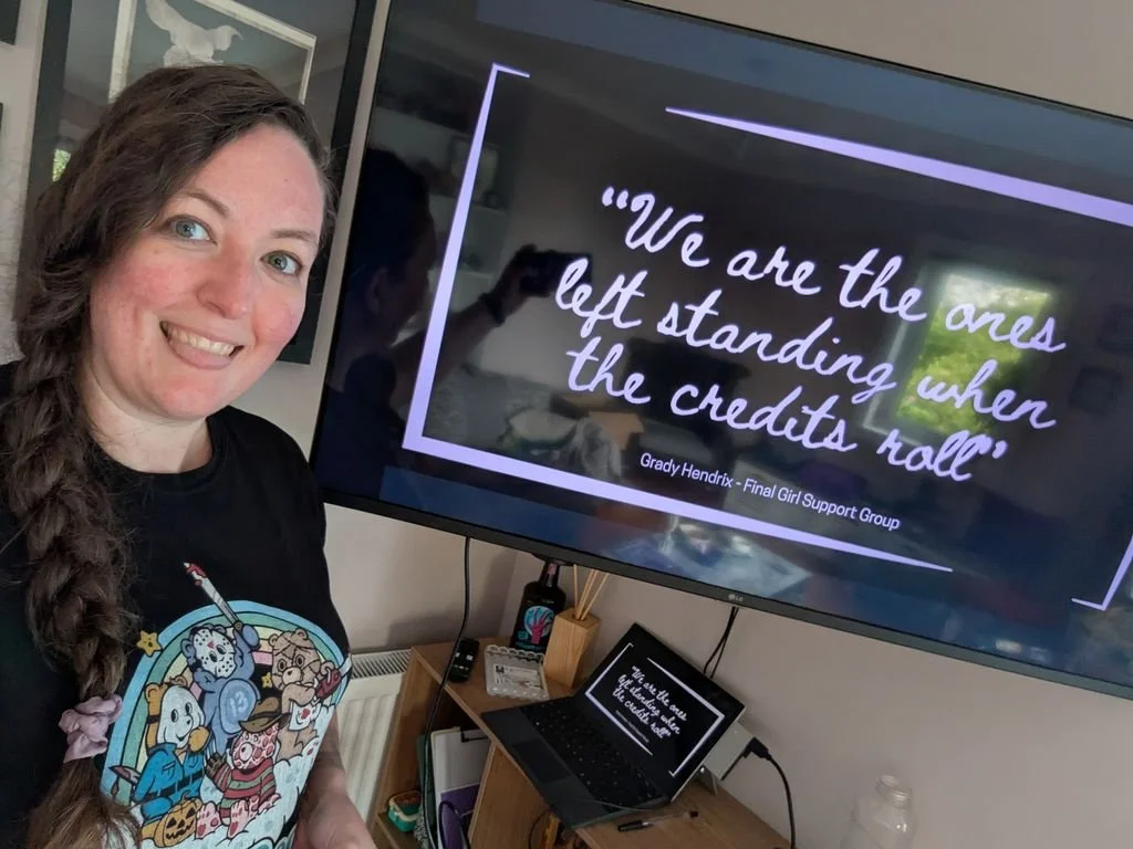 A woman with long brown hair and a braid is smiling at the camera, standing next to a TV screen displaying a quote. The quote reads: 'We are the ones left standing when the credits roll.' The TV is placed on a wooden desk with a laptop, remote, and writing supplies, and a smaller screen below shows the same quote.