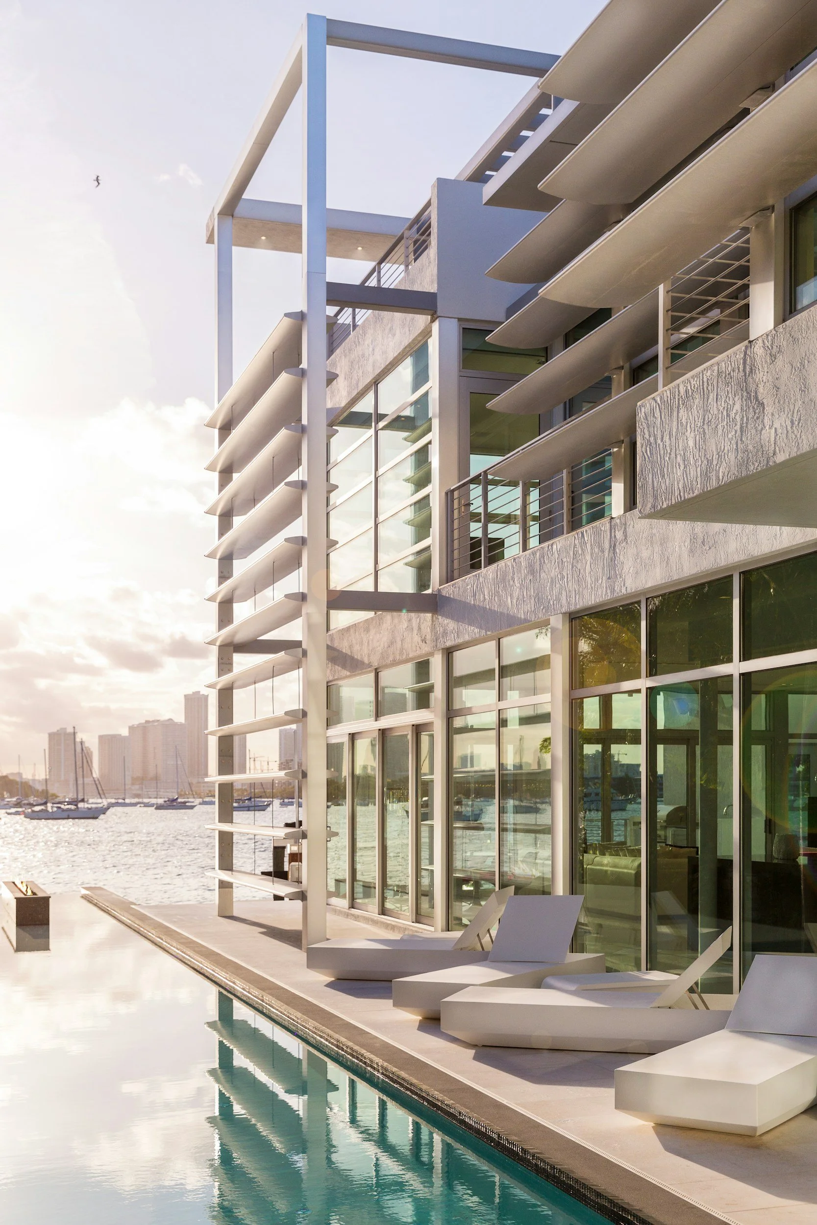 Modern waterfront house with large windows, architectural detailing, poolside loungers, and an infinity pool overlooking a city skyline and marina in the background.