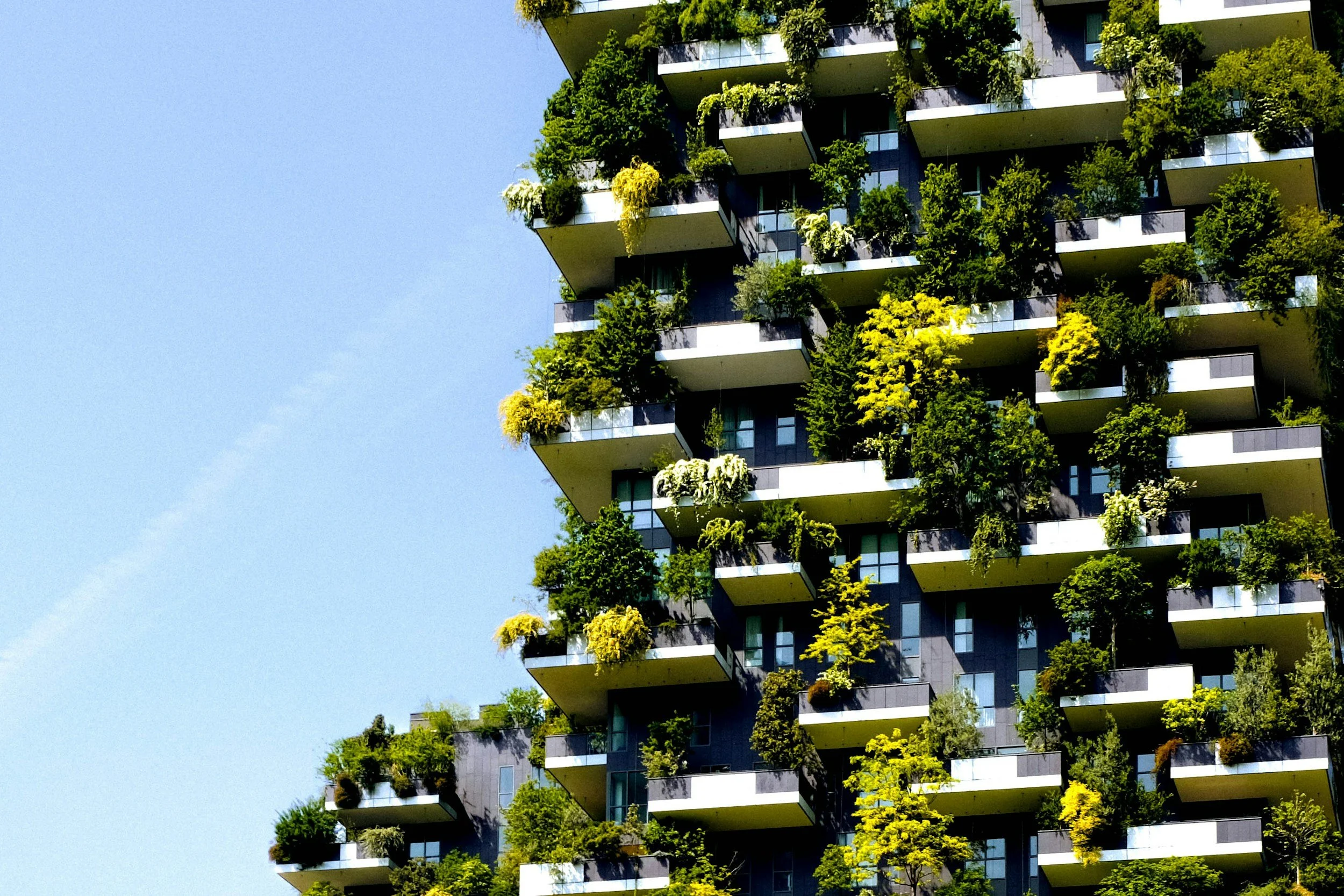 A modern residential building with balconies covered in lush greenery and plants against a clear blue sky.