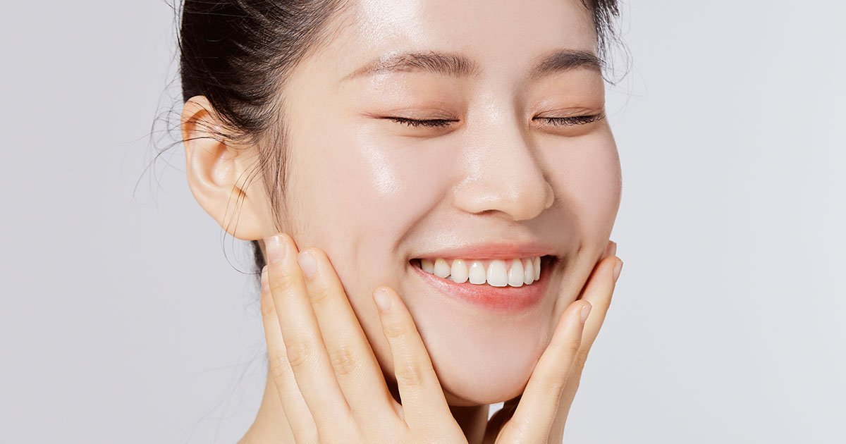 A woman with clear skin smiling with eyes closed, lightly touching her face with both hands.