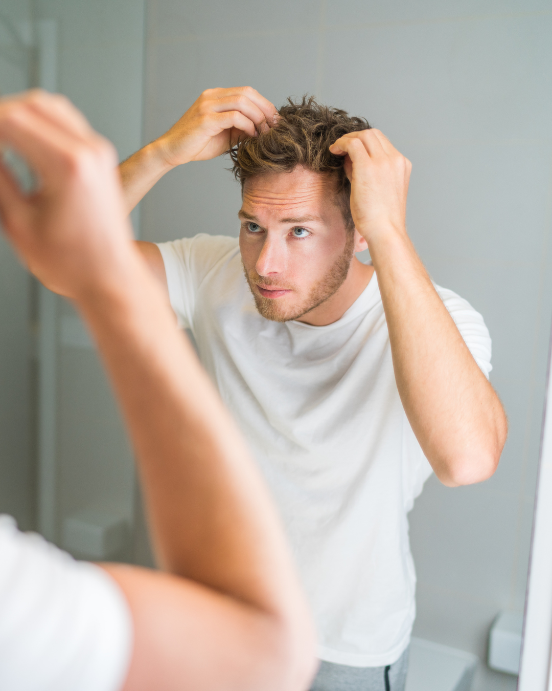 A young man with light skin and short, wavy hair looking in the mirror while fixing or styling his hair.