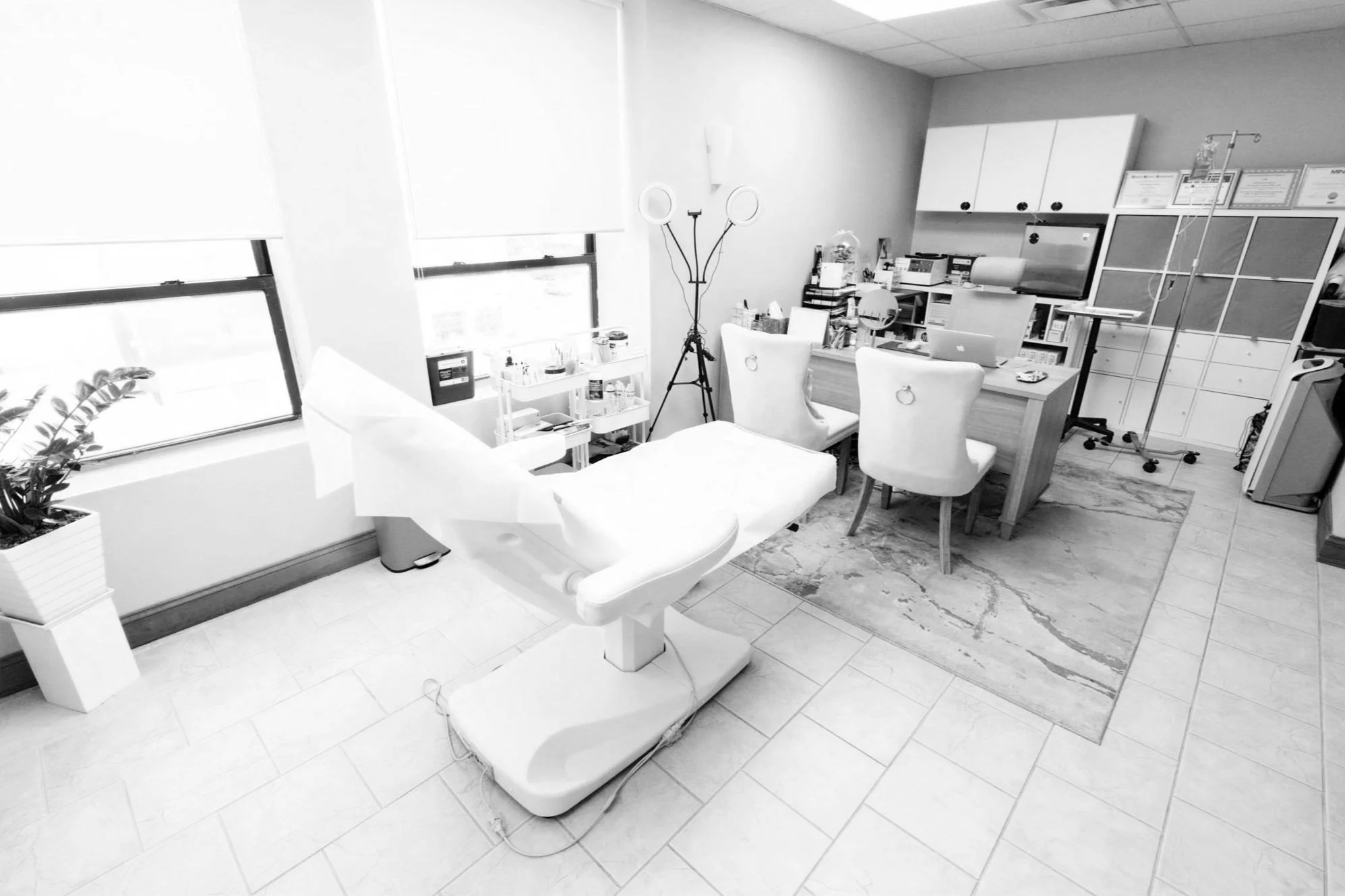 Medical examination room with an exam table, computer, and office chairs, medical equipment, and supplies; large windows with blinds, and various medical certificates on the wall.