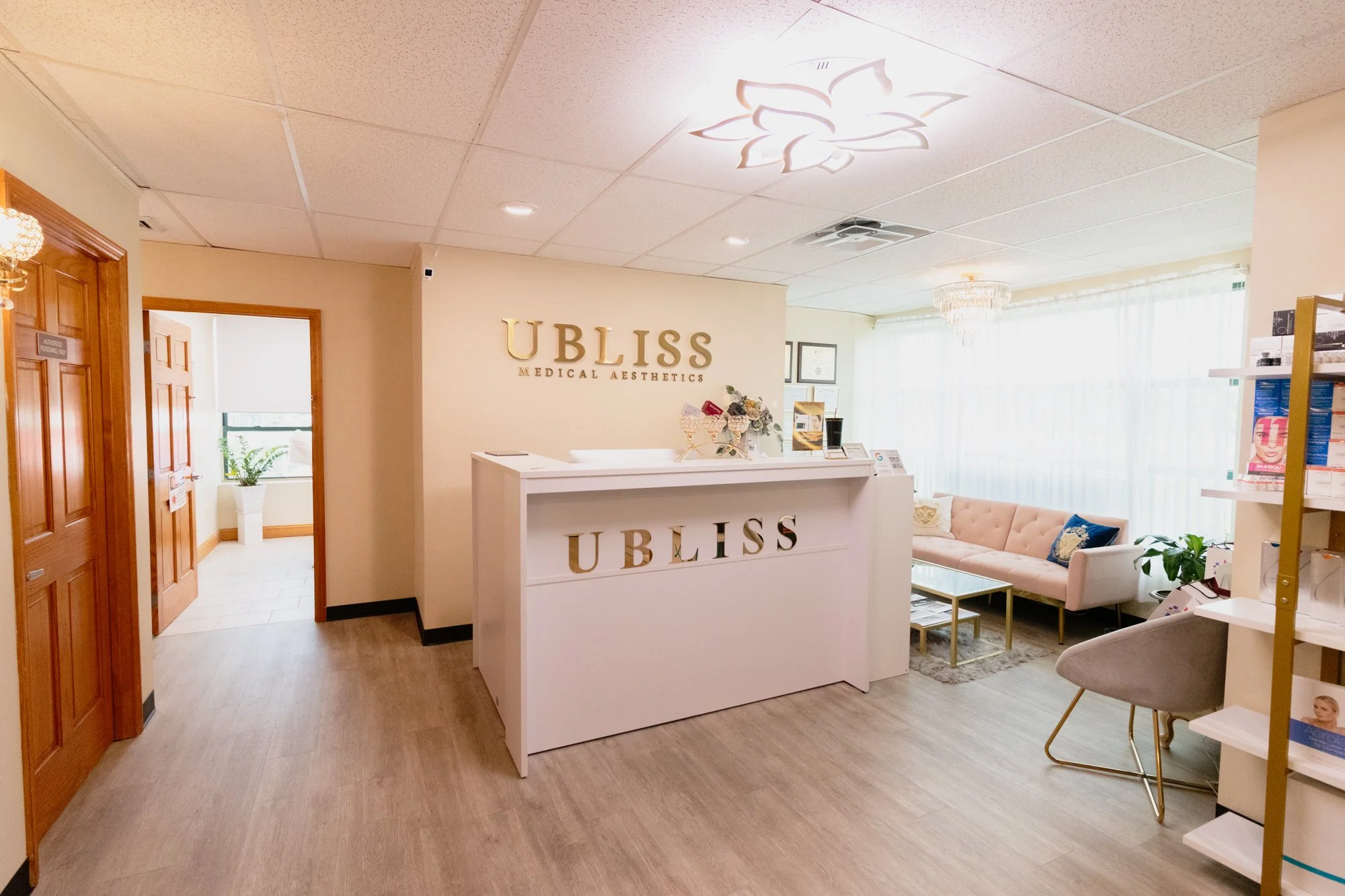 Reception area of a clinic named UBLISS MEDICAL AESTHETICS with a white counter, gold lettering, and a seating area with a pink sofa and a glass coffee table.