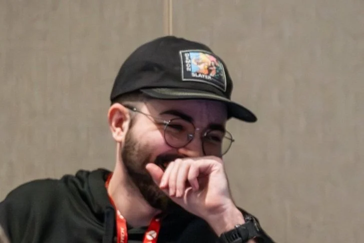 Man with glasses and a beard wearing a black cap, laughing and covering his mouth with his hand.