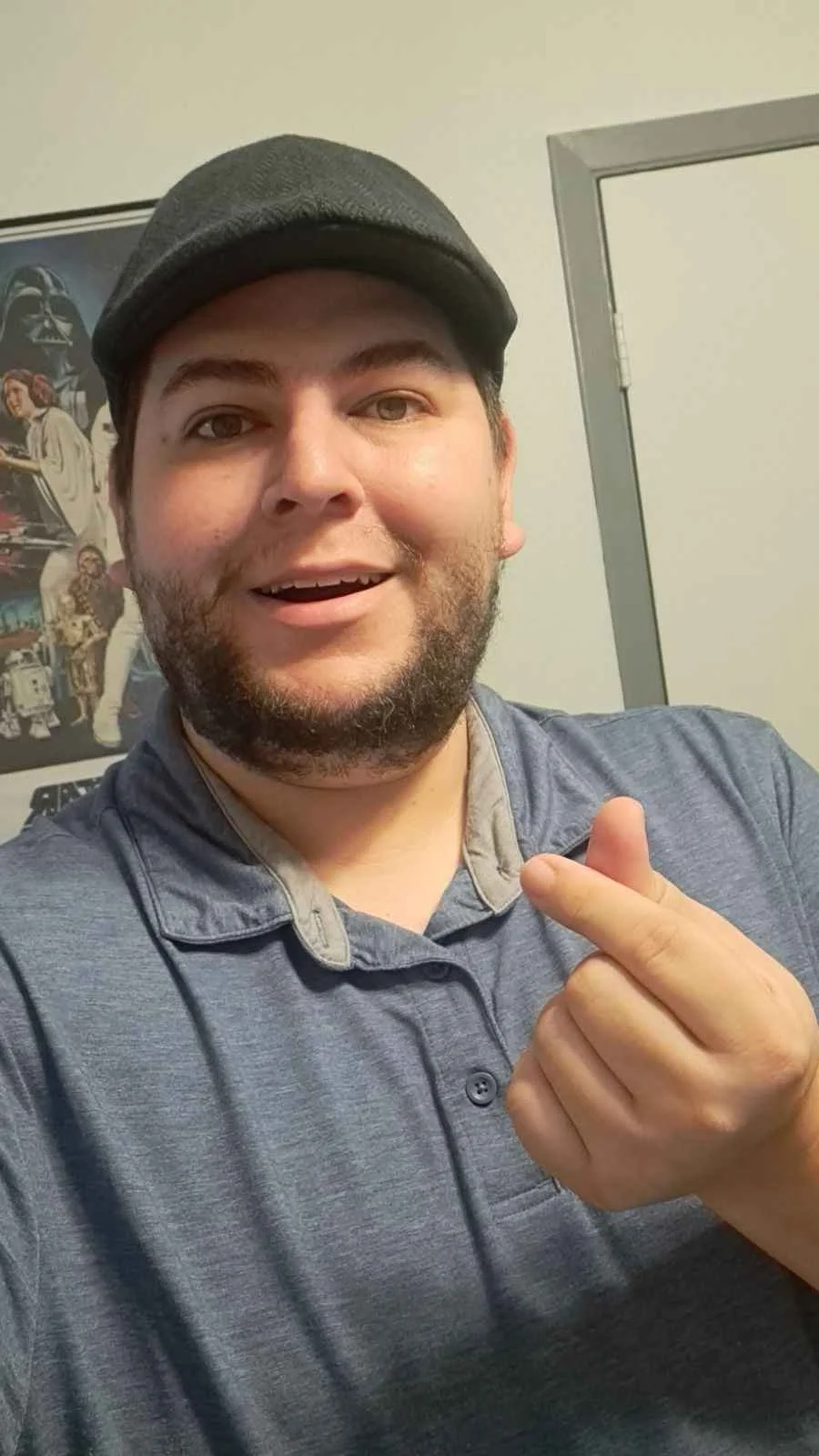 A man with a beard and black cap making a finger heart gesture in front of a Star Wars poster.