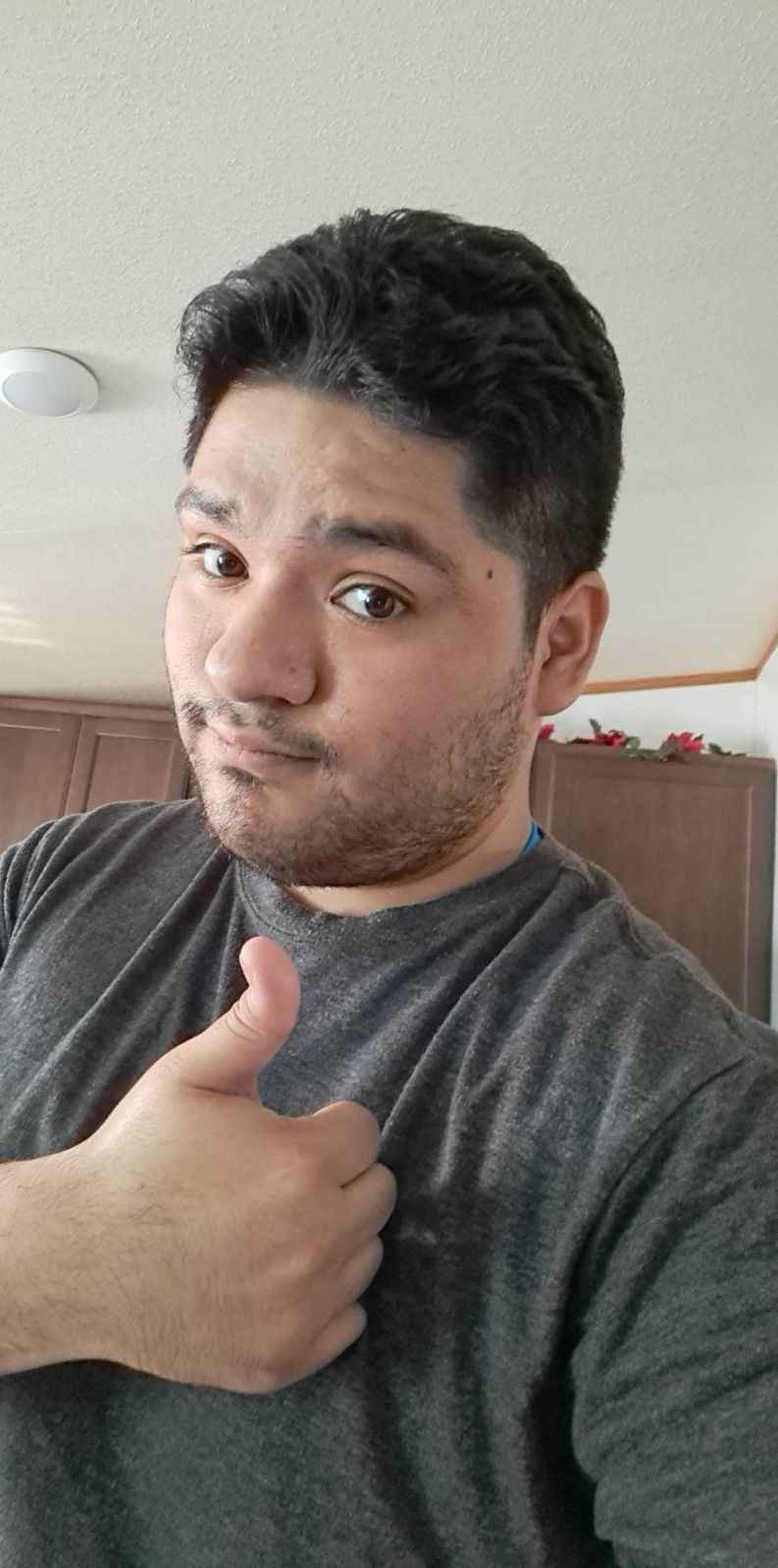 A young man with dark hair giving a thumbs up and winking at the camera inside a home, with wooden cabinets and holiday decorations in the background.