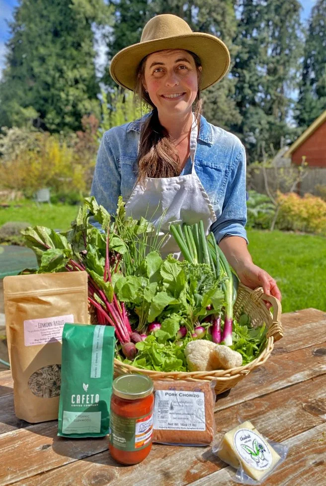 A woman with a wide-brimmed hat and denim shirt standing behind a basket of fresh vegetables, including radishes, green onions, and lettuce, on a wooden table outdoors with trees and a house in the background.