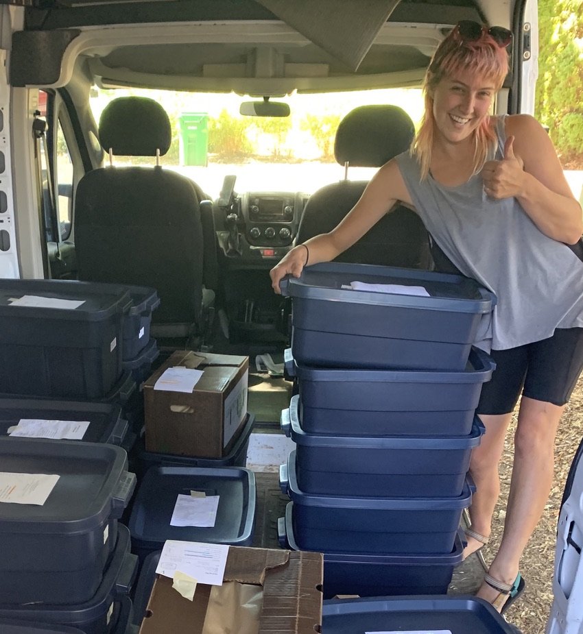 A woman with pink hair giving a thumbs-up while leaning on large labeled plastic storage bins inside a vehicle, surrounded by more bins and paperwork.