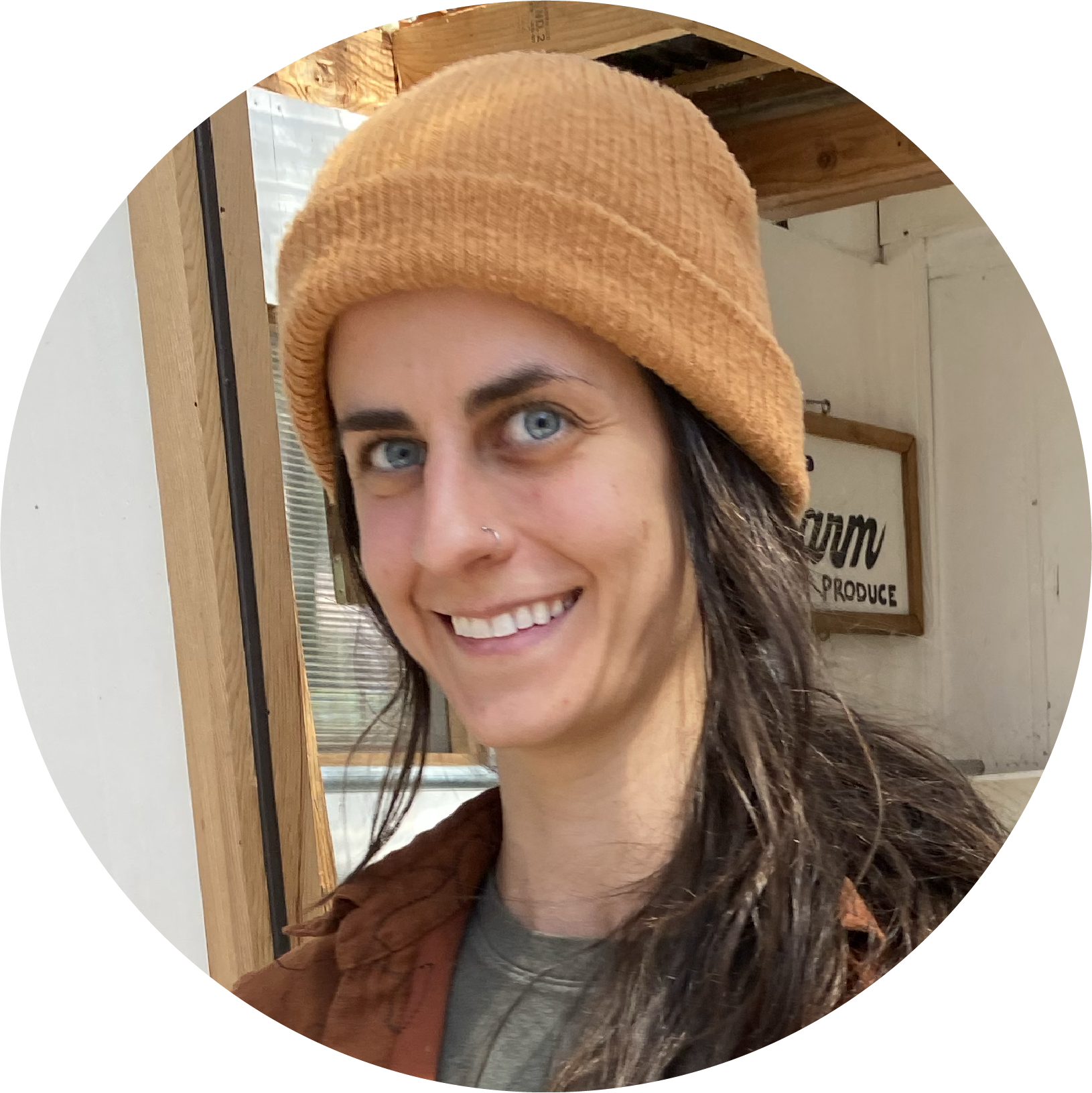 A woman smiling, with blue eyes, dark hair, and wearing a brown beanie hat and a brown jacket, standing inside a building with wooden elements in the background.