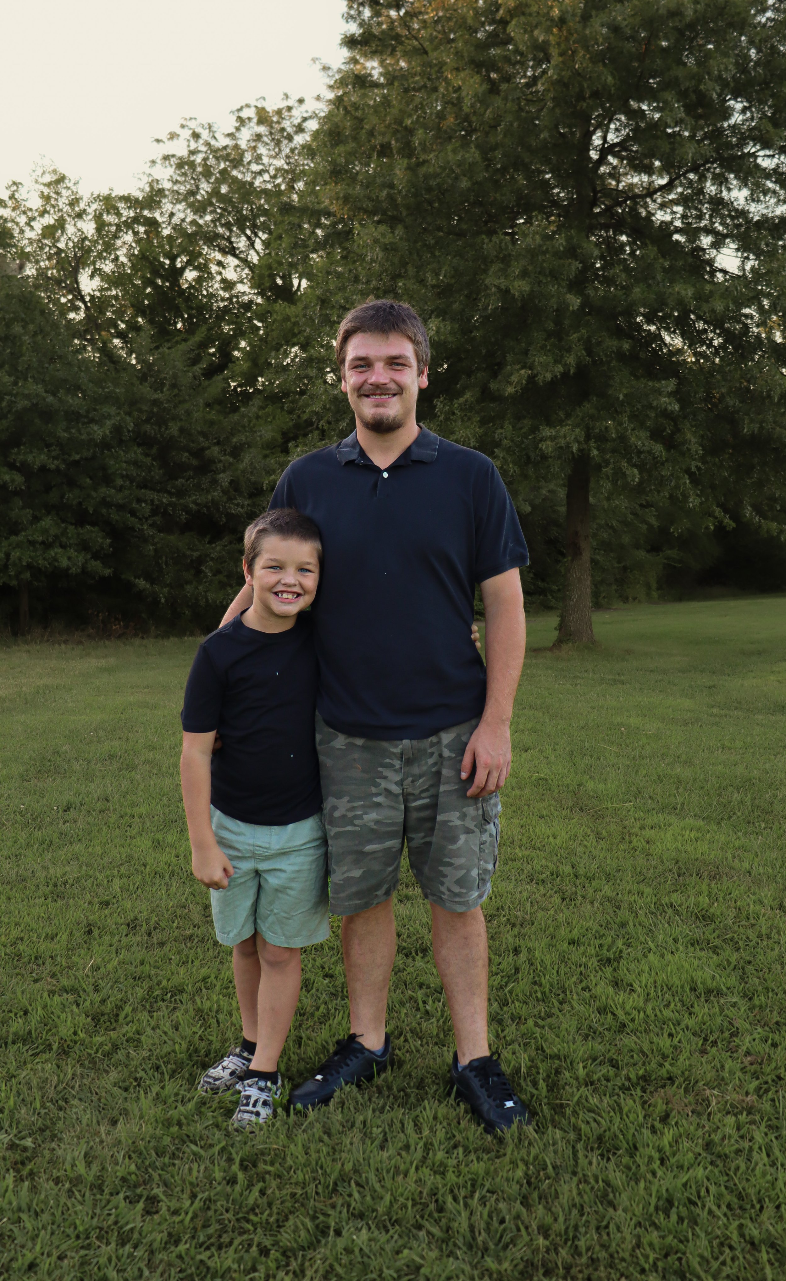 A man and boy standing together outdoors on a grassy field with trees in the background, both smiling at the camera.