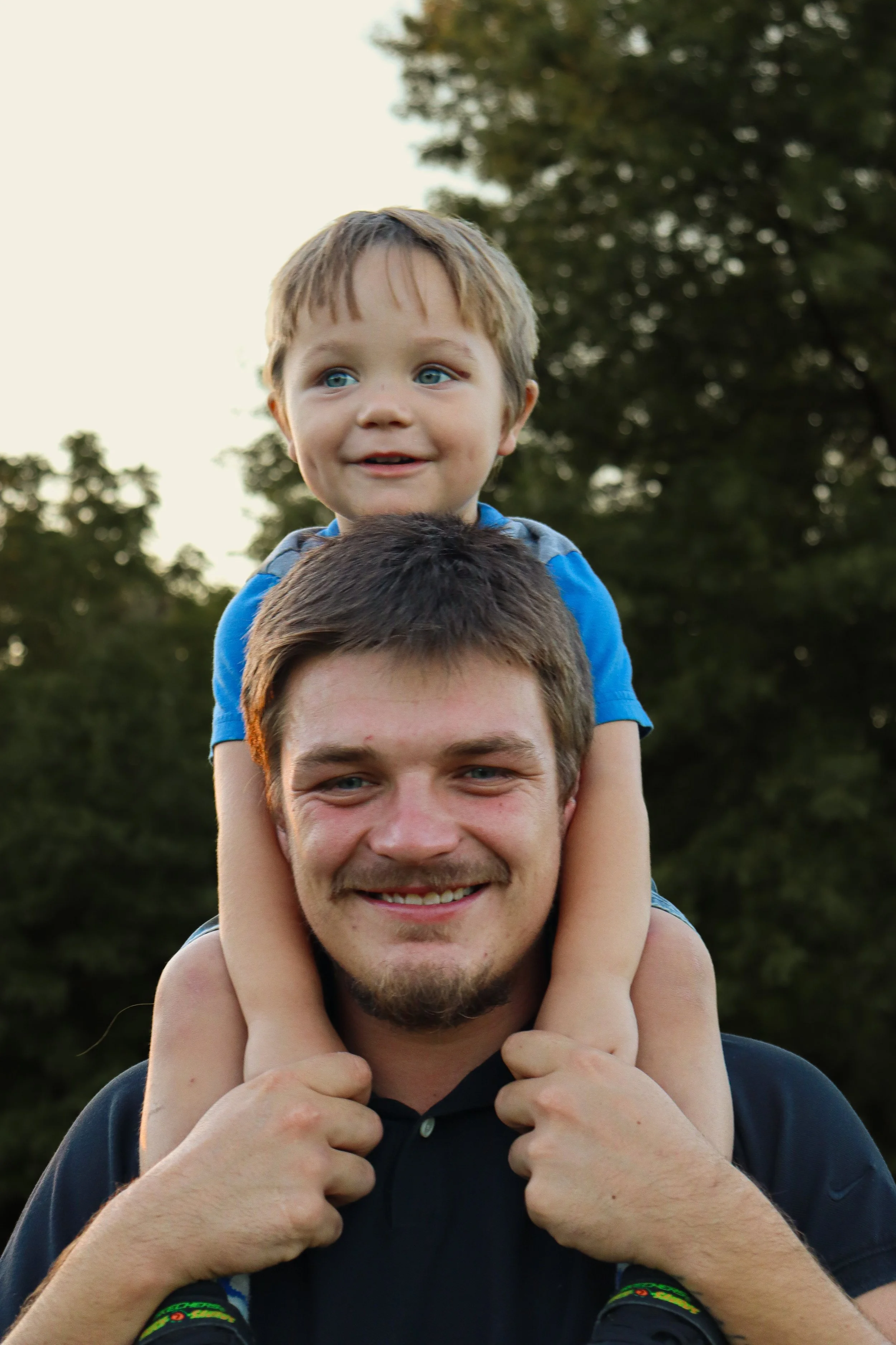 A man with short brown hair and a beard carrying a young boy with blond hair on his shoulders, both smiling outdoors with trees in the background.