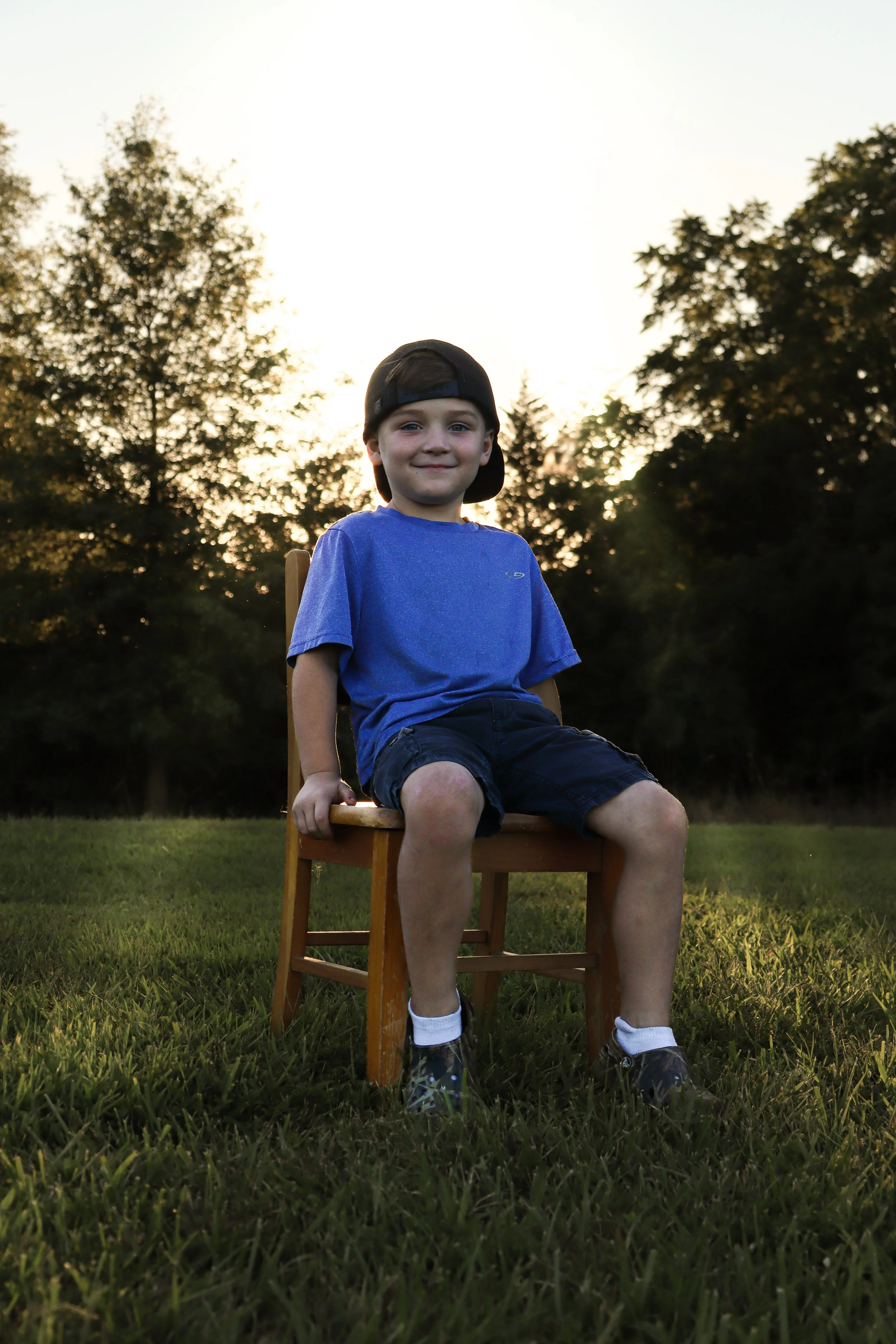 A young boy sitting on a wooden chair outdoors during sunset, wearing a blue t-shirt, black shorts, a black cap backwards, and sneakers with white socks.