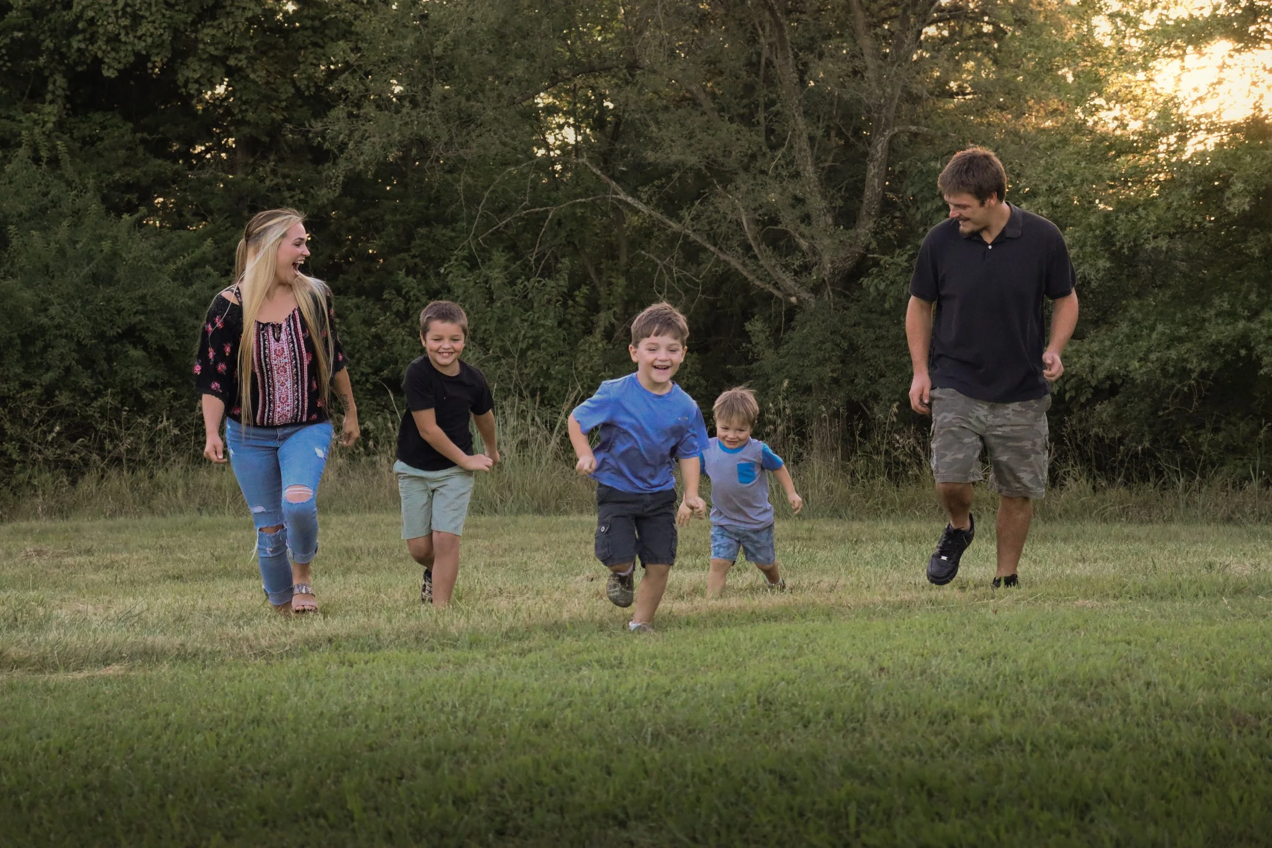 A family of six, including two adults and four children, running and playing in a grassy field during sunset with trees in the background.