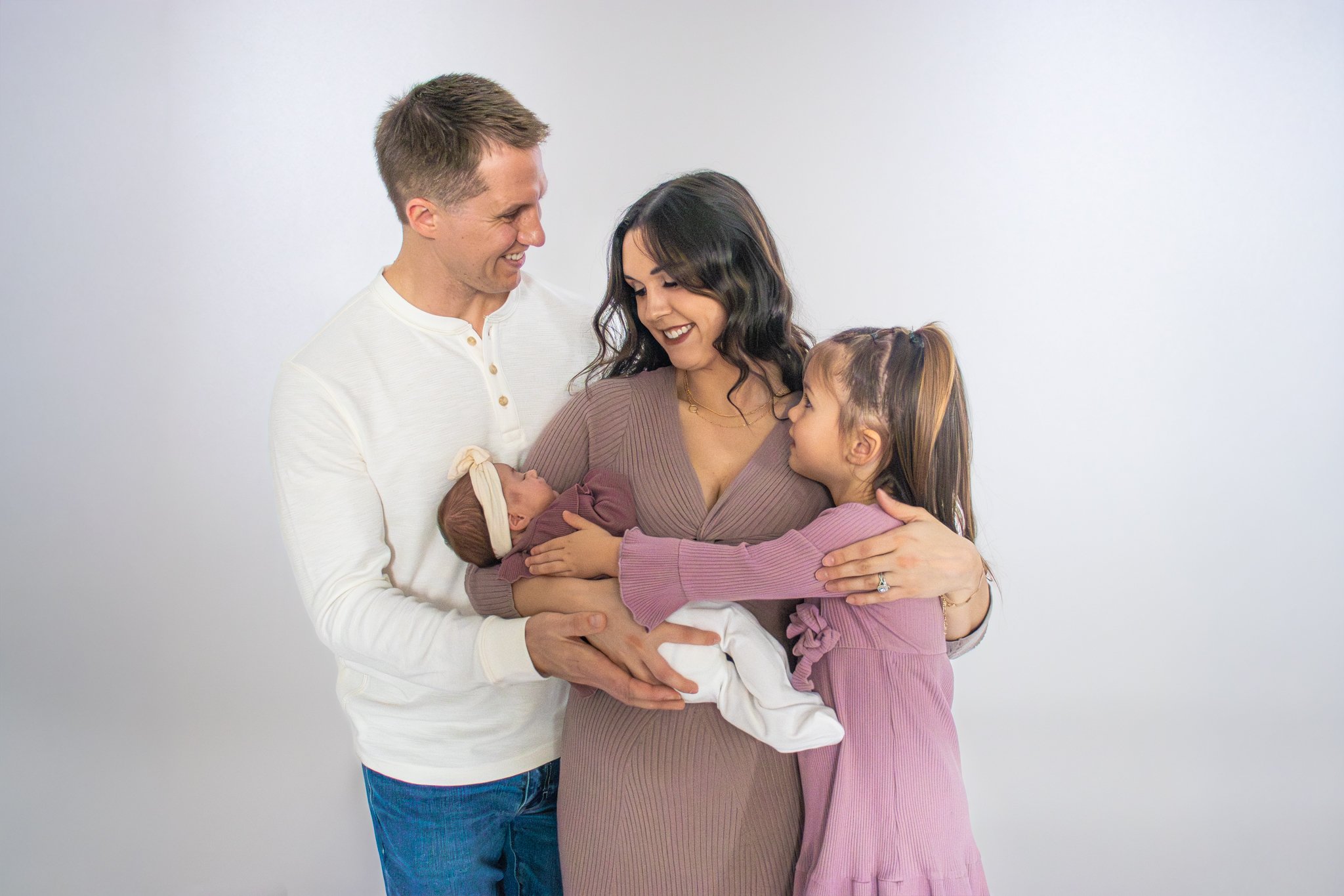 A family of four standing together, with the mother holding a newborn baby, and the father and daughter looking at the baby, all smiling warmly.